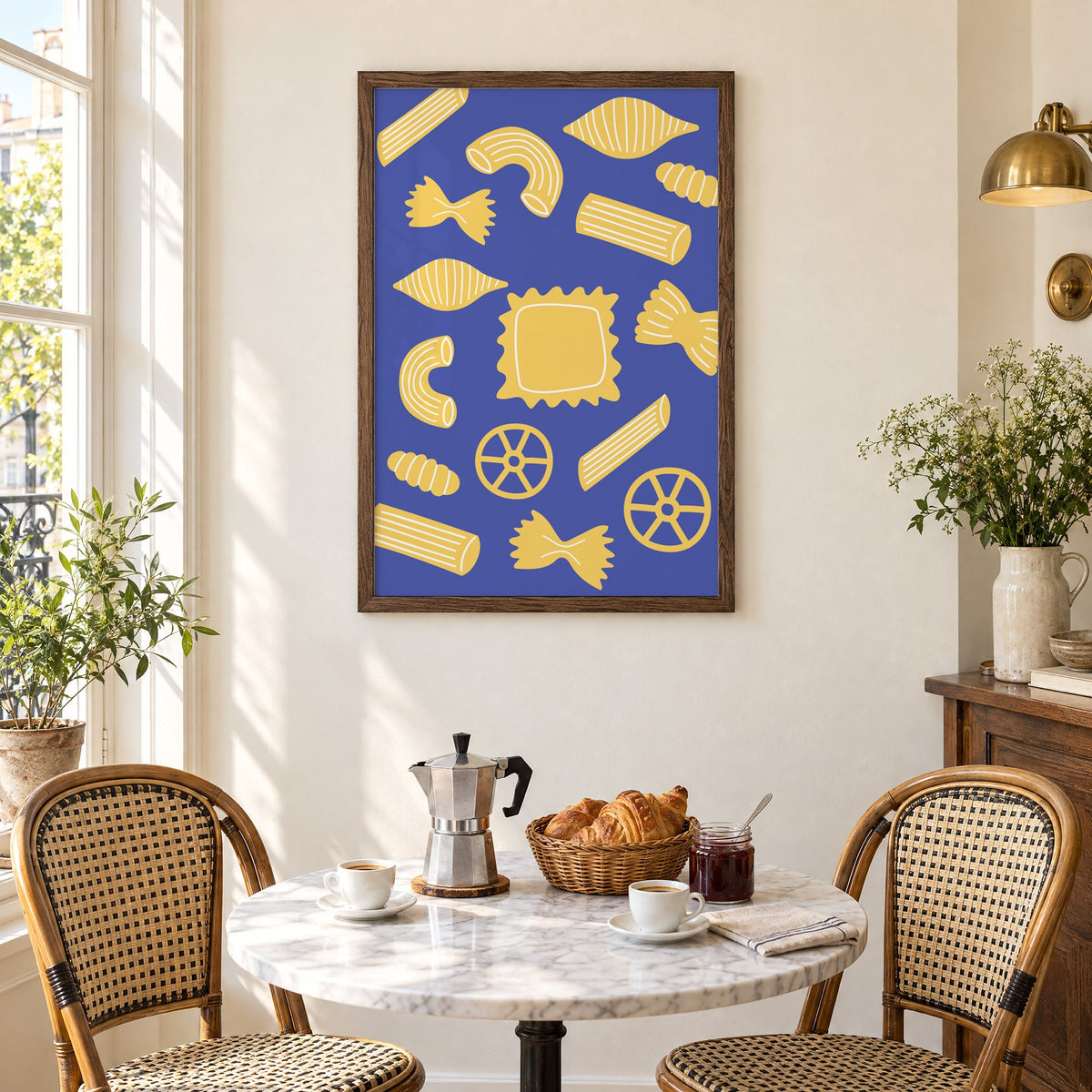 Yellow pasta shapes pattern on blue print in a walnut frame above a marble bistro table with croissants and rattan chairs