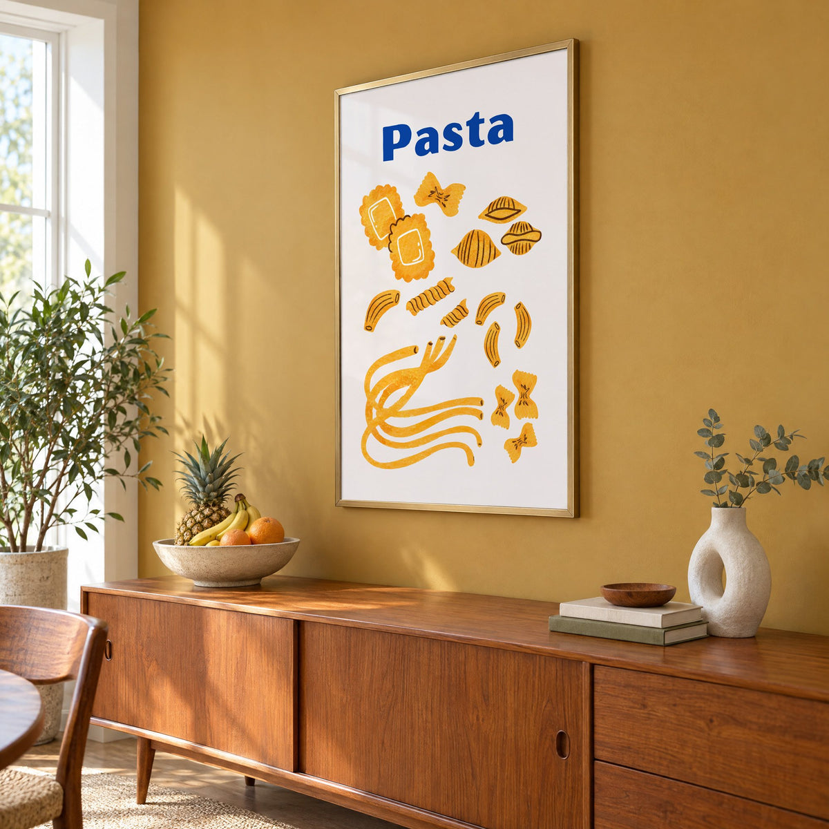Pasta blue title watercolor pasta shapes print in a brass frame on a mustard wall above a teak sideboard with a fruit bowl, seen at an angle