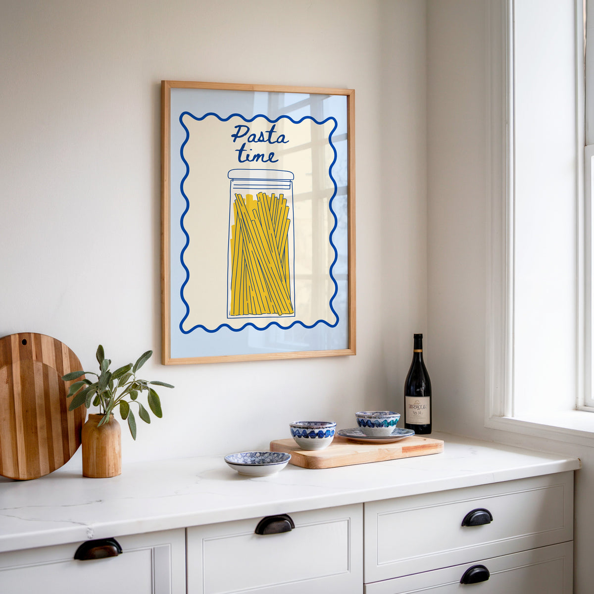 Pasta Time jar print in an oak frame above a white sideboard with a bottle of wine