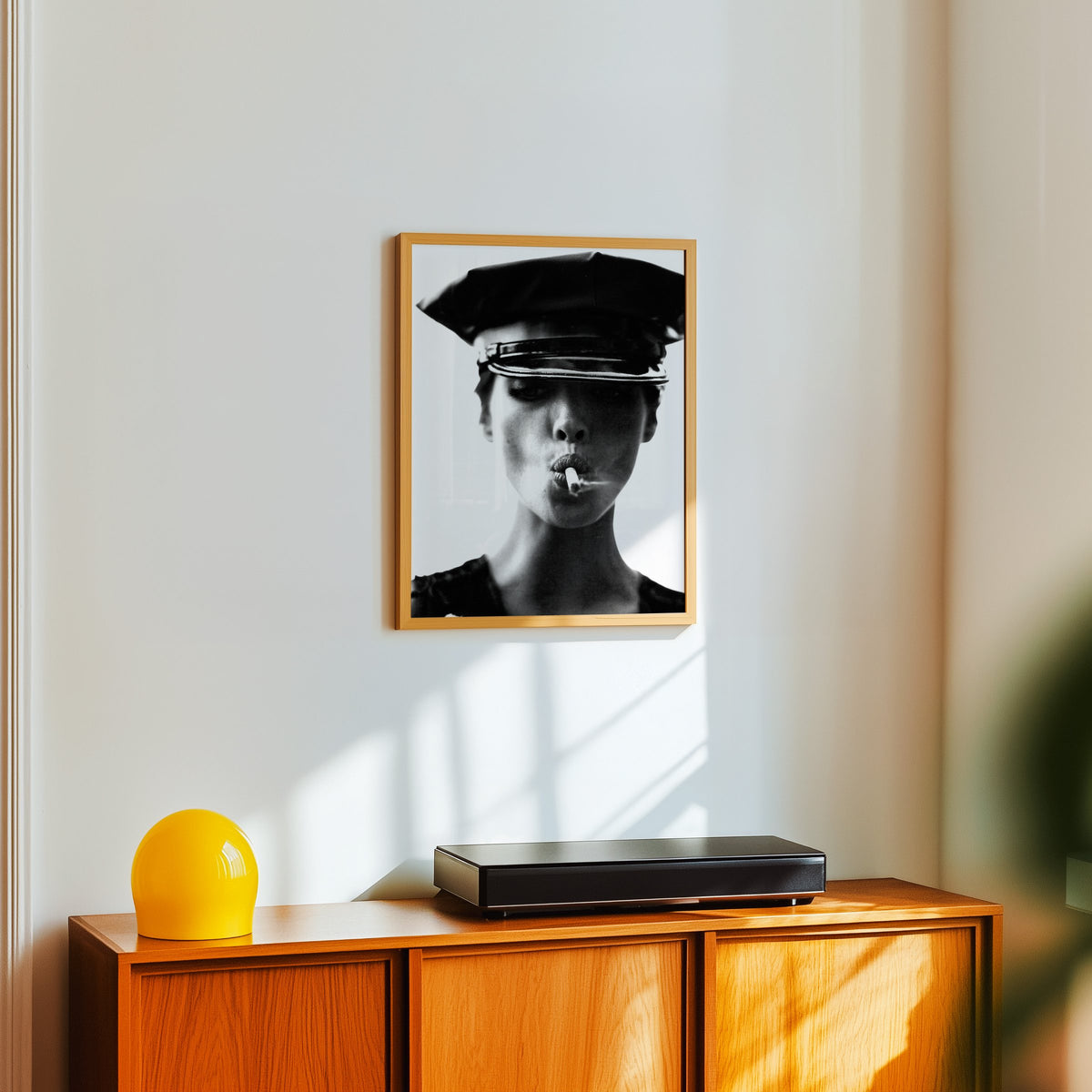 Close portrait under glossy leather cap print in an oak frame above a teak sideboard with a yellow glass lamp