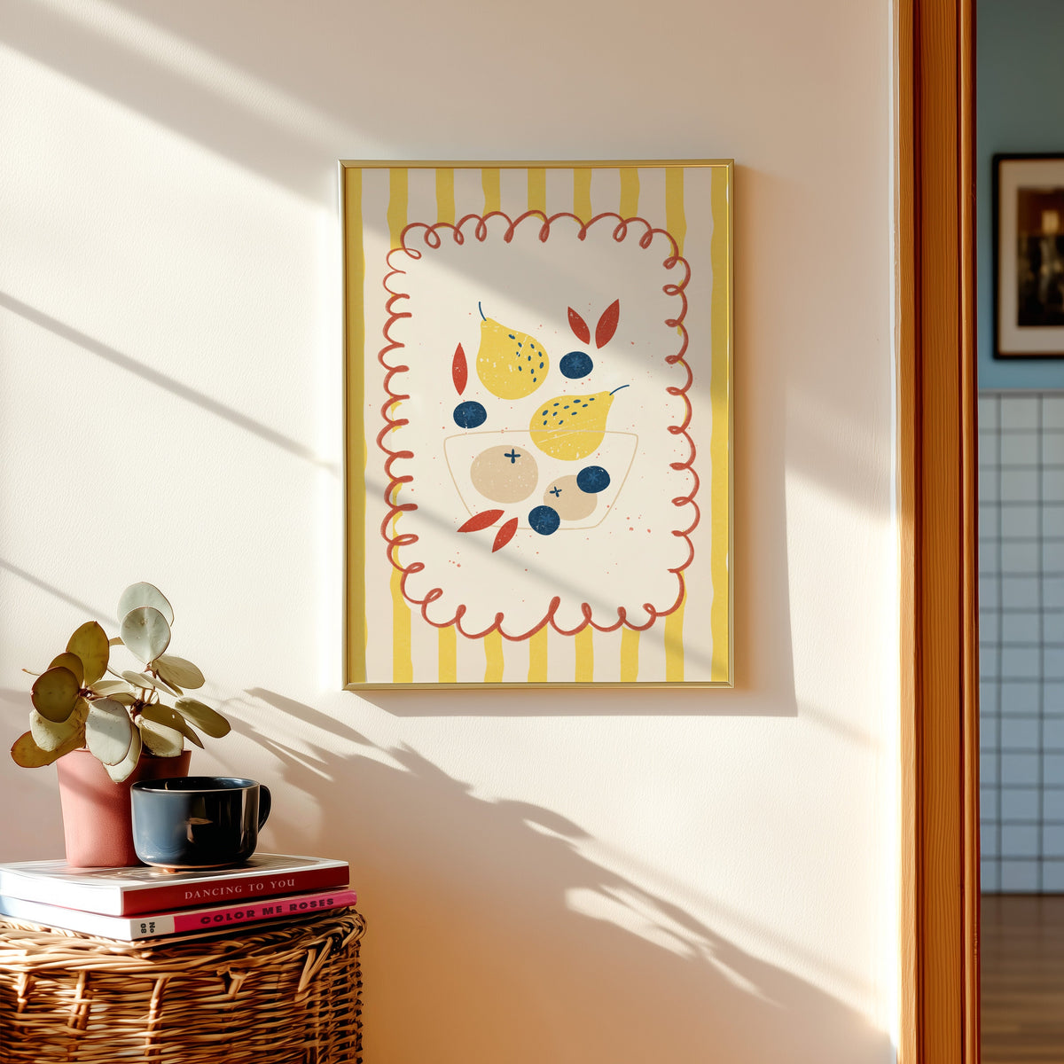 Pears and blueberries print in a gold frame above a basket of books and a pilea plant