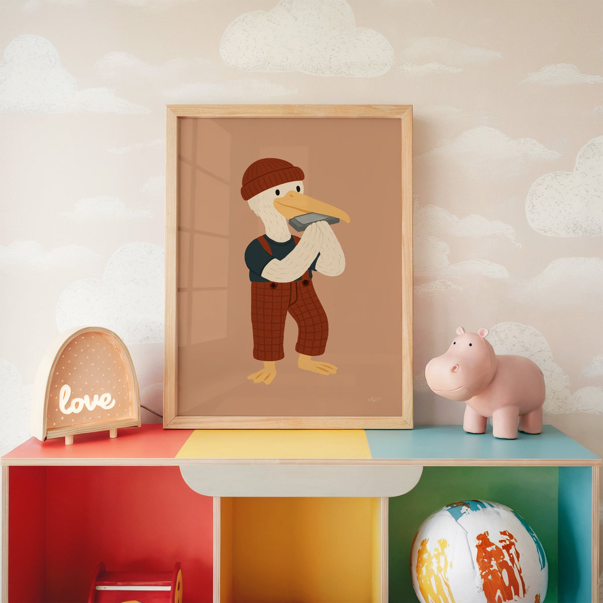 Pelican with harmonica and beanie nursery print in an oak frame on a rainbow toy shelf against cloud wallpaper