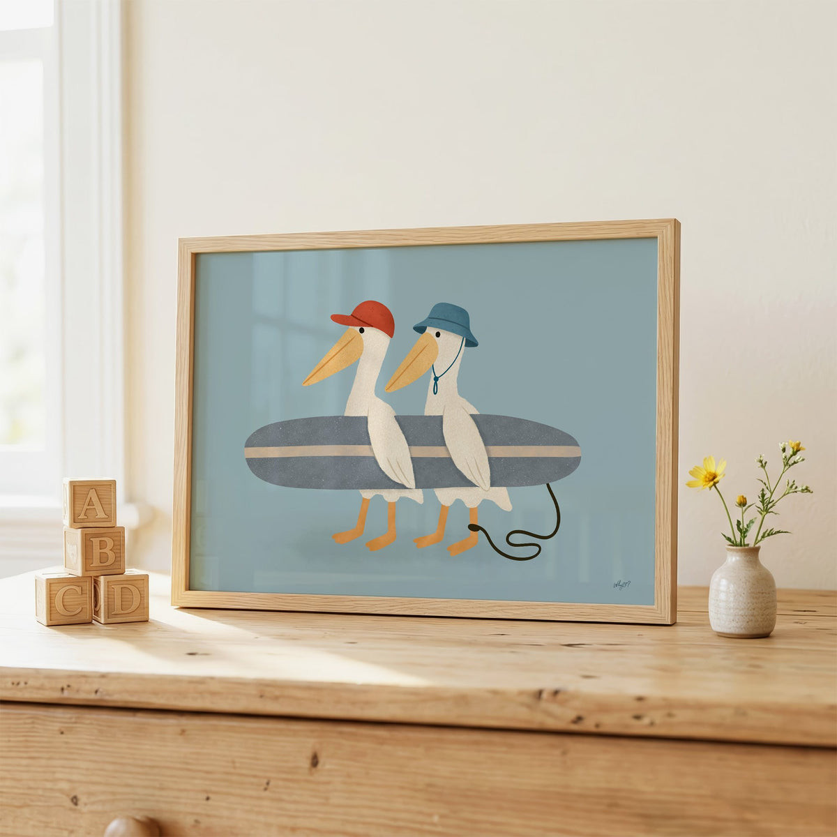 Two pelicans carrying surfboard nursery print in an oak frame on a wooden dresser beside ABC blocks