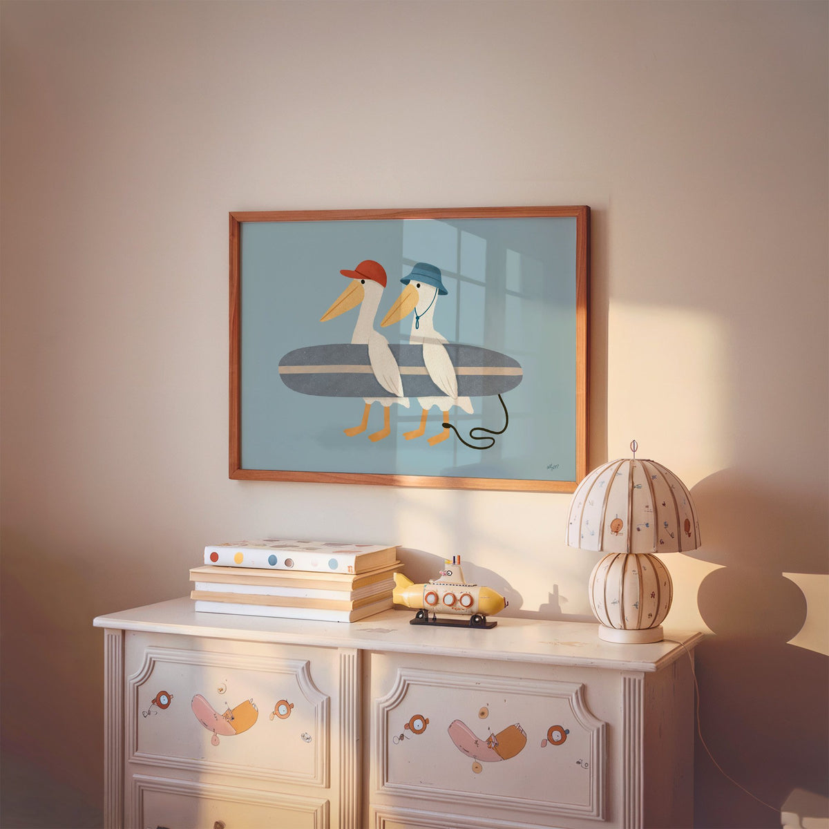 Two pelicans carrying surfboard nursery print in a cherry wood frame above a painted dresser and lamp
