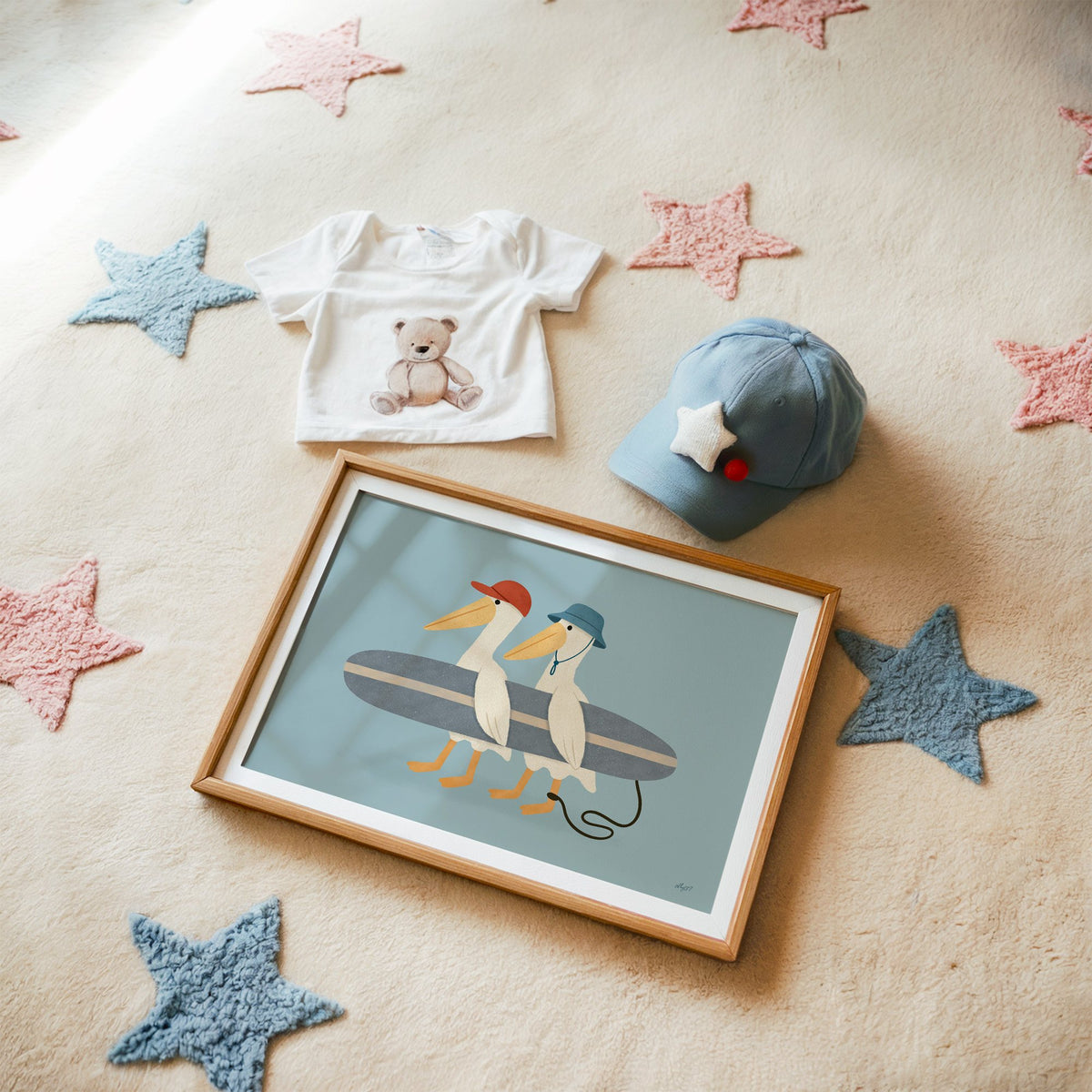 Two pelicans carrying surfboard nursery print in a matted oak frame on a star rug with a baby tee