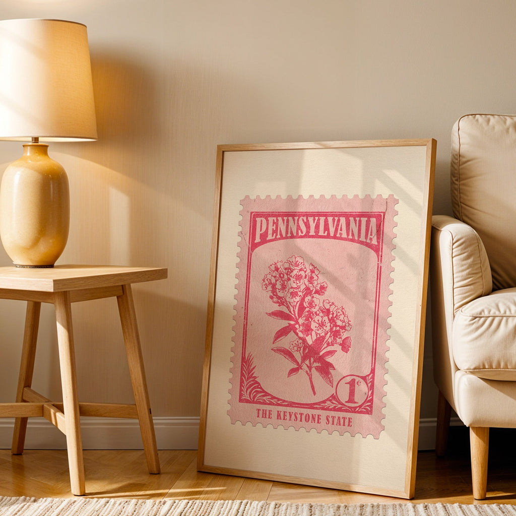 Pennsylvania stamp print in a wood frame resting on the floor beside a beige armchair and a yellow lamp