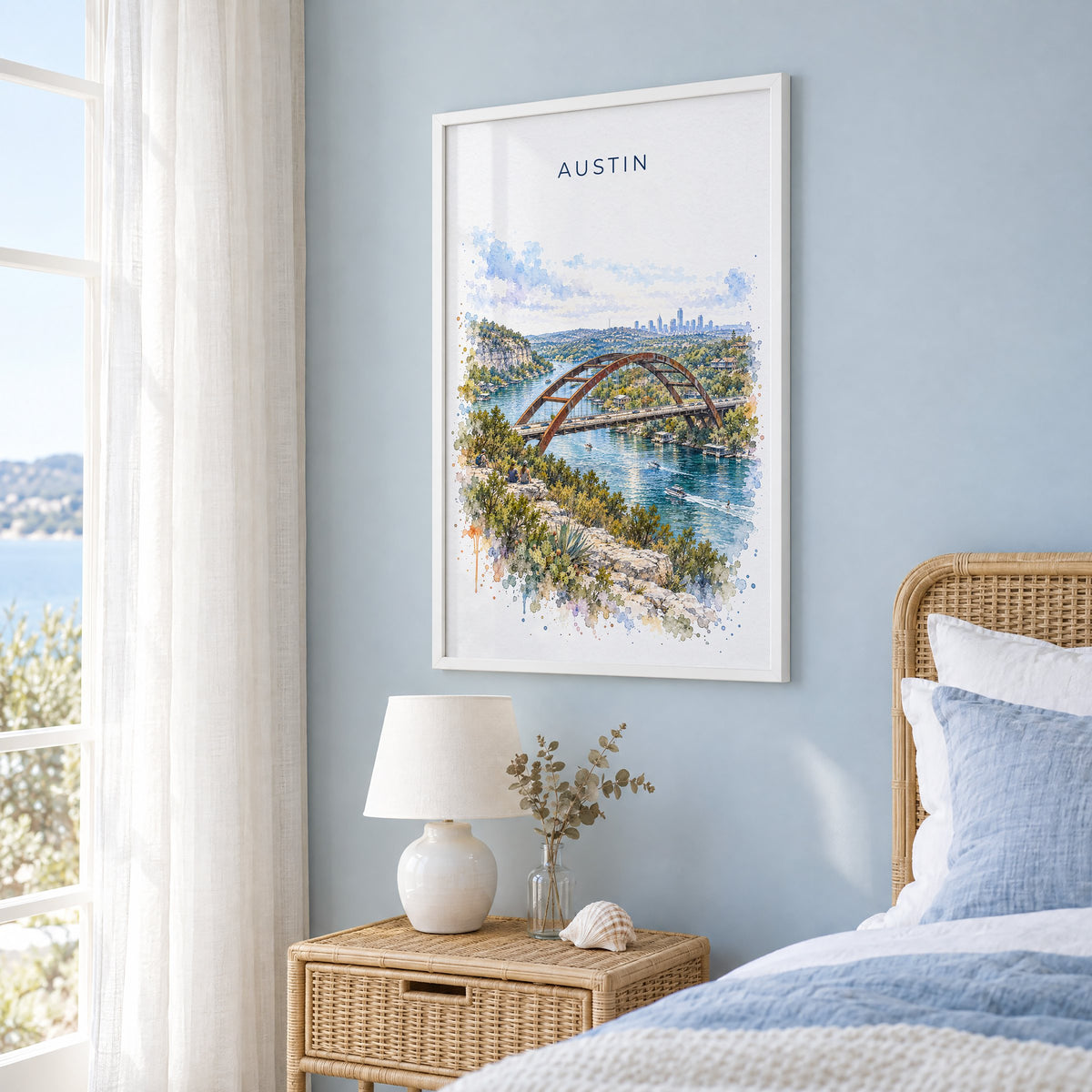 Pennybacker Bridge Austin watercolor print in a white frame on a pale blue wall beside a rattan bed in a coastal bedroom