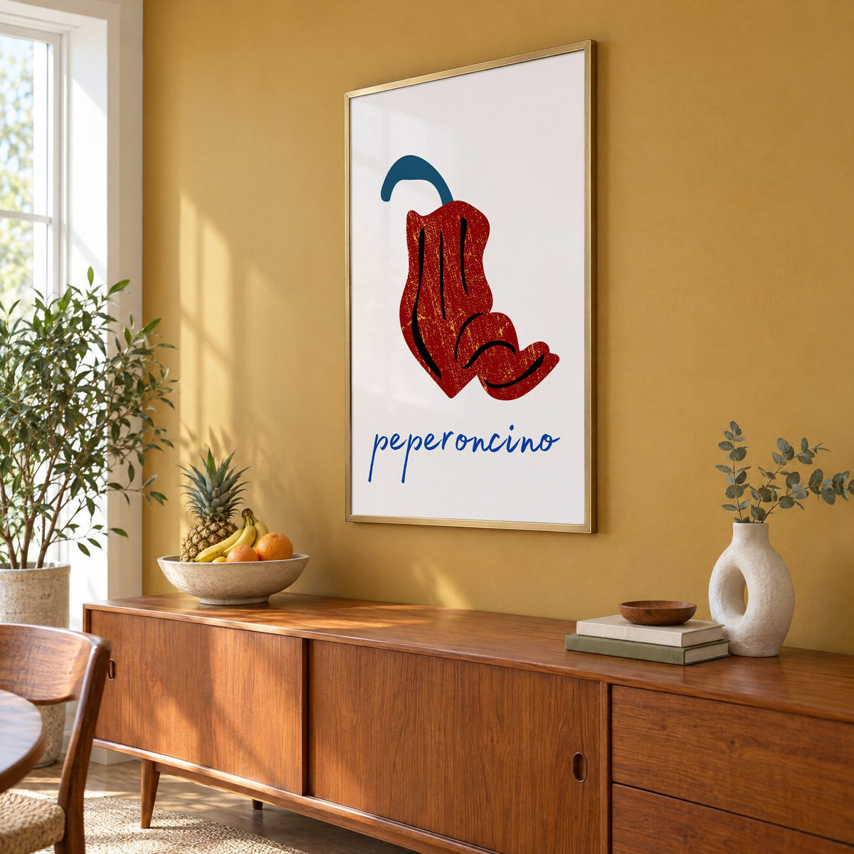 peperoncino red chili grunge print in a brass frame on a mustard wall above a teak sideboard with a fruit bowl, seen at an angle