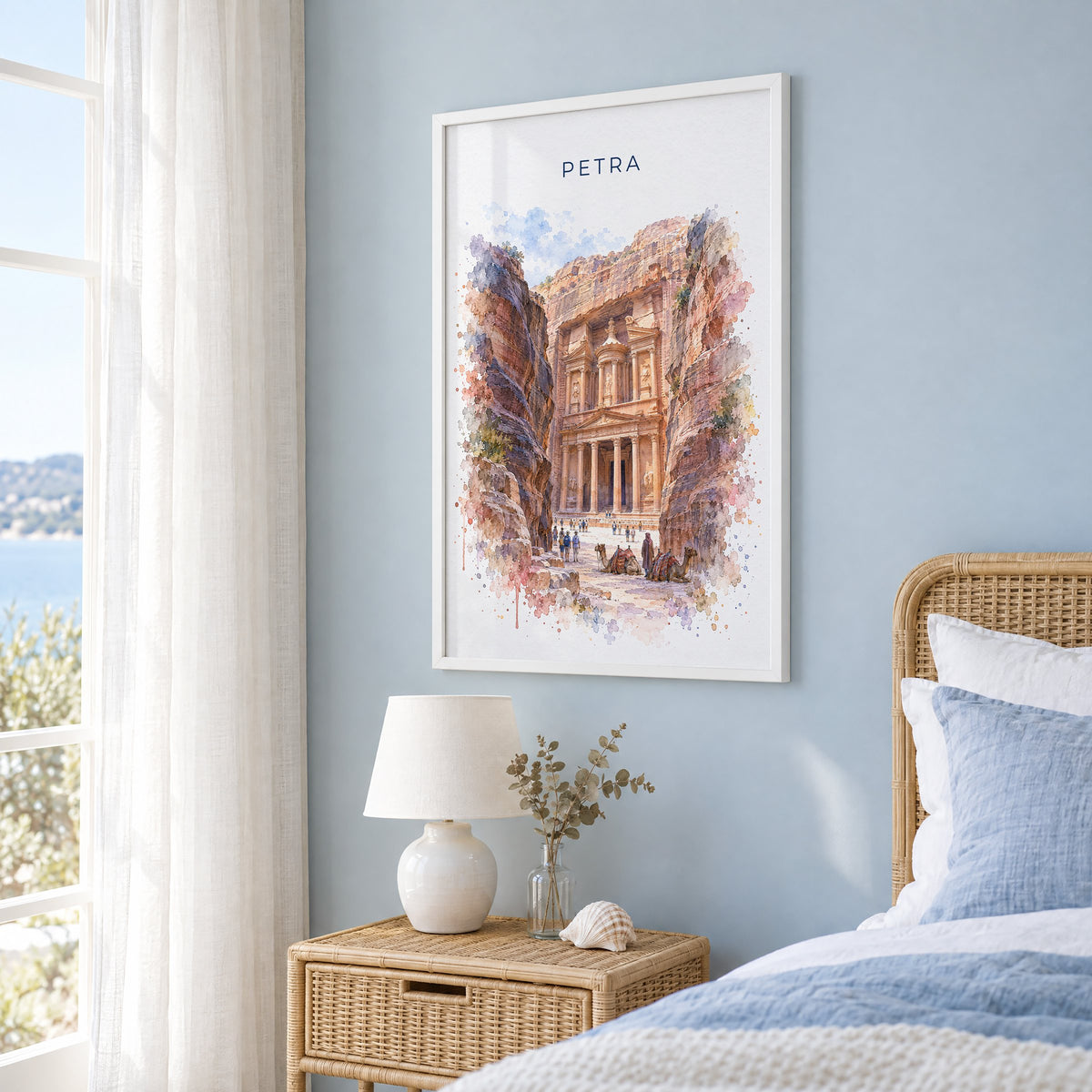 Petra watercolor print in a white frame on a pale blue wall beside a rattan bed in a coastal bedroom