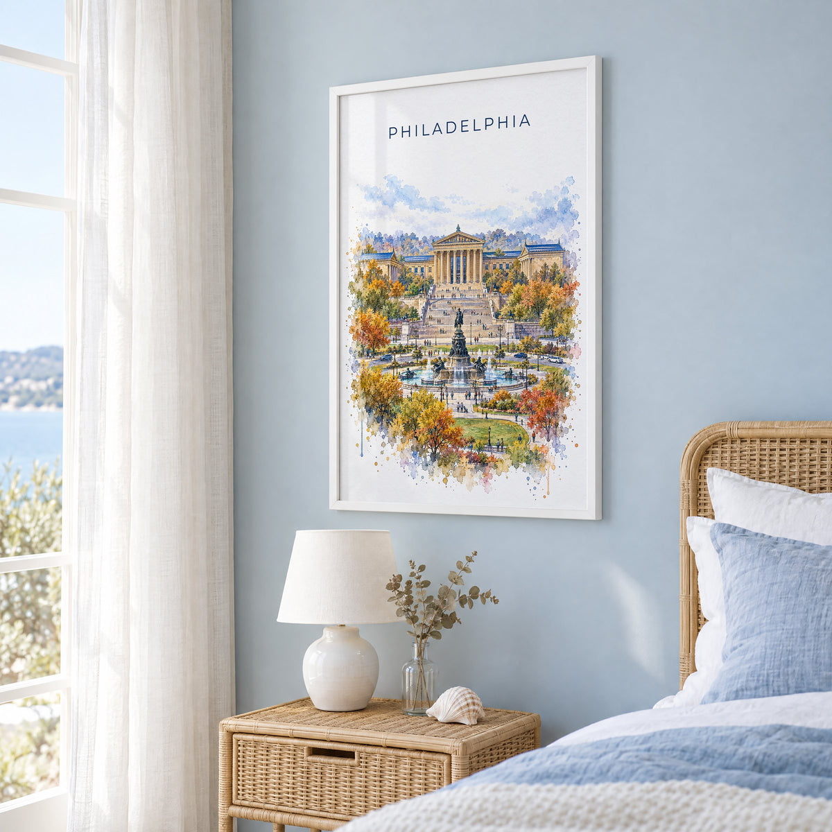 Philadelphia Museum of Art watercolor print in a white frame on a pale blue wall beside a rattan bed in a coastal bedroom