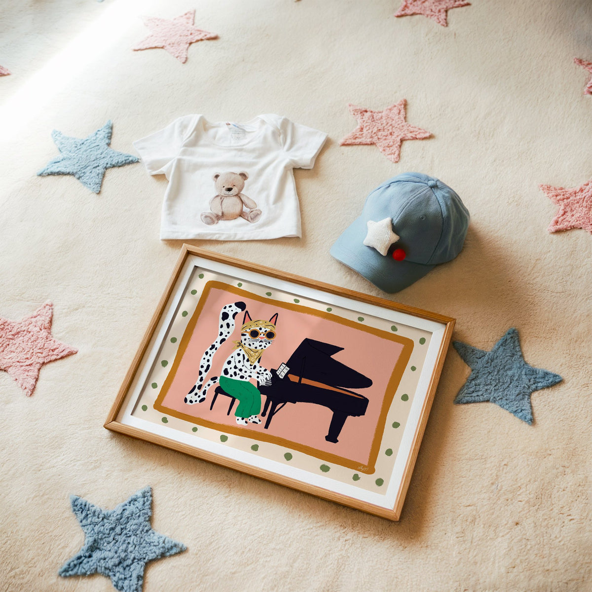 Spotted cat playing grand piano nursery print in a matted oak frame on a star rug with a baby tee