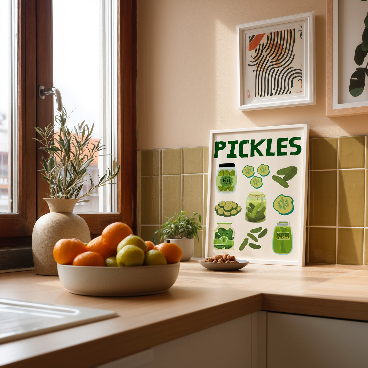 Pickles jars and slices chart print in a white frame leaning against olive green kitchen tiles beside a bowl of citrus