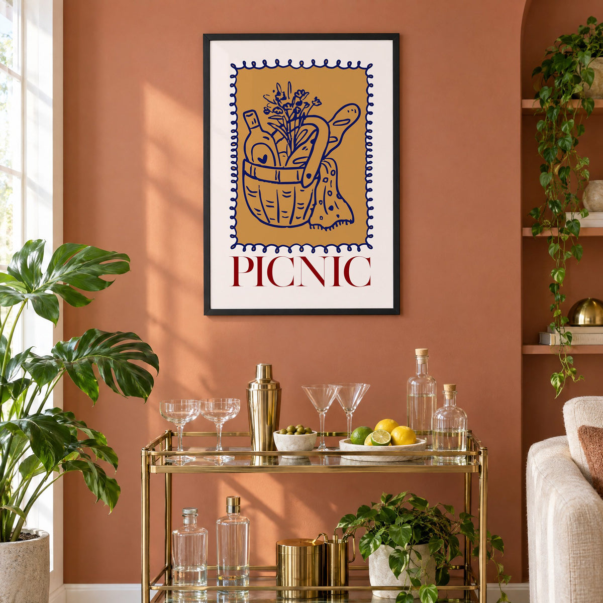 PICNIC basket wine and baguette stamp print in a black frame on a terracotta wall above a brass bar cart with coupes and citrus