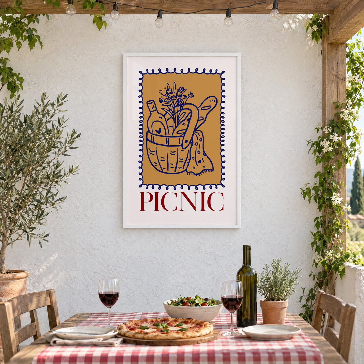 PICNIC basket wine and baguette stamp print in a white frame on a stucco wall above a checked-cloth table with pizza and red wine