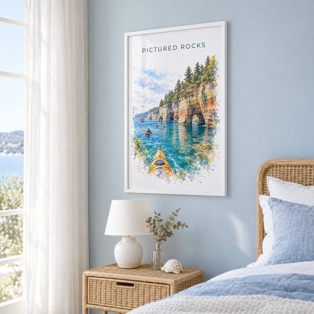Pictured Rocks watercolor print in a white frame on a pale blue wall beside a rattan bed in a coastal bedroom