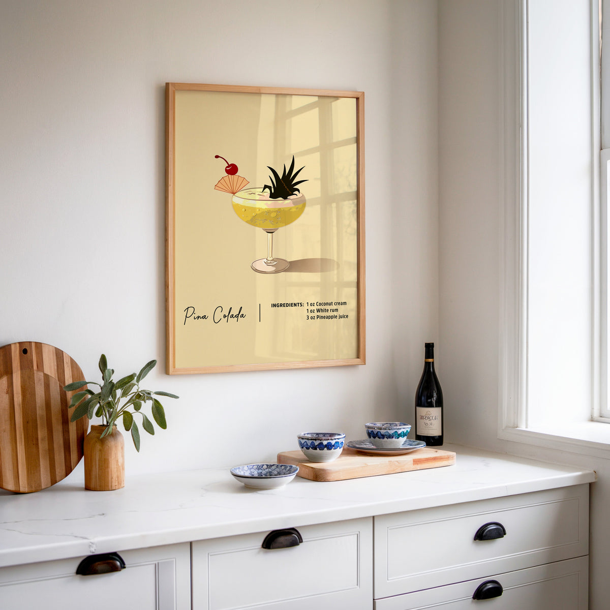 Pina colada recipe print in an oak frame above a white sideboard with a bottle of wine