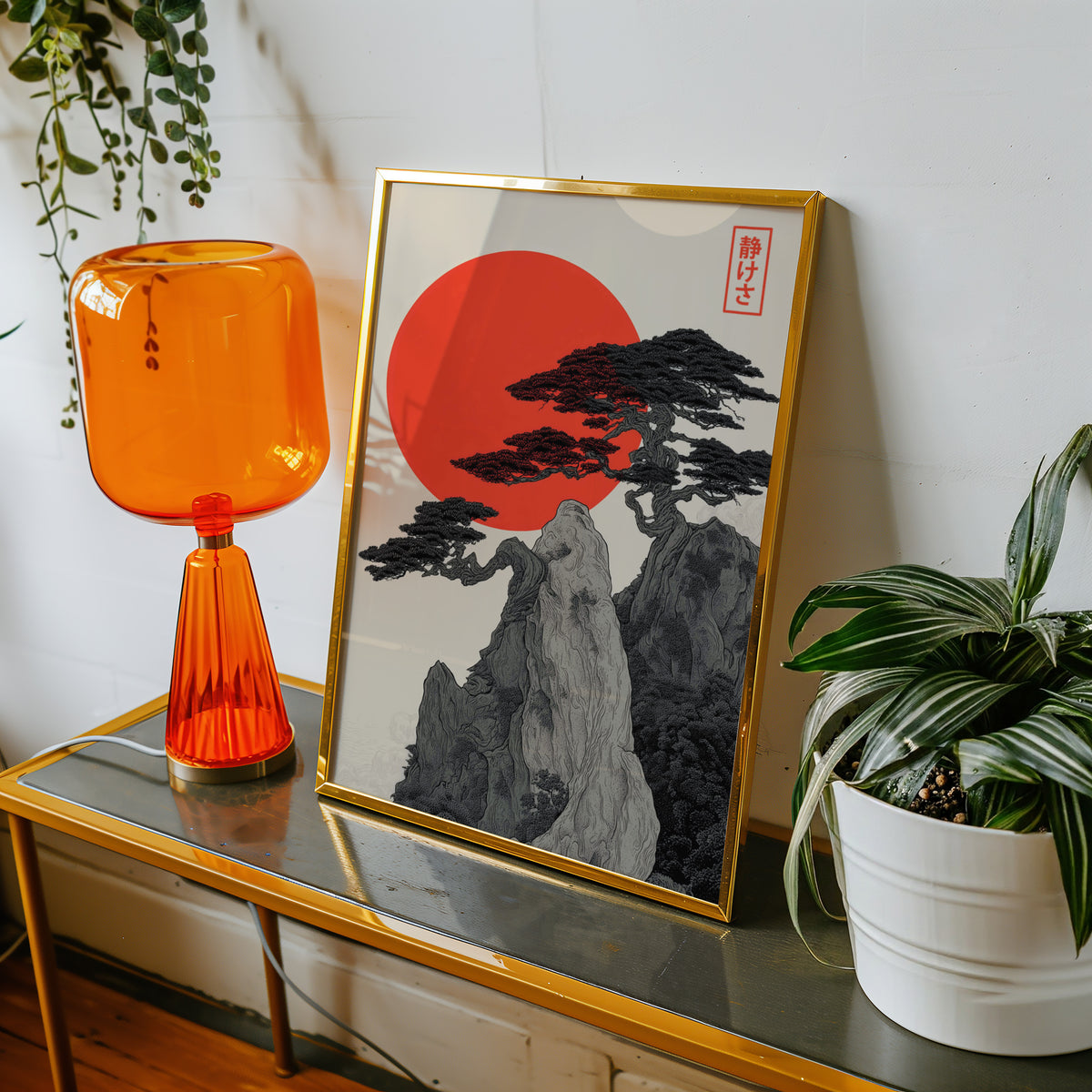 Ink pine cliff with red sun print shown in a gold frame on a console beside an orange glass lamp
