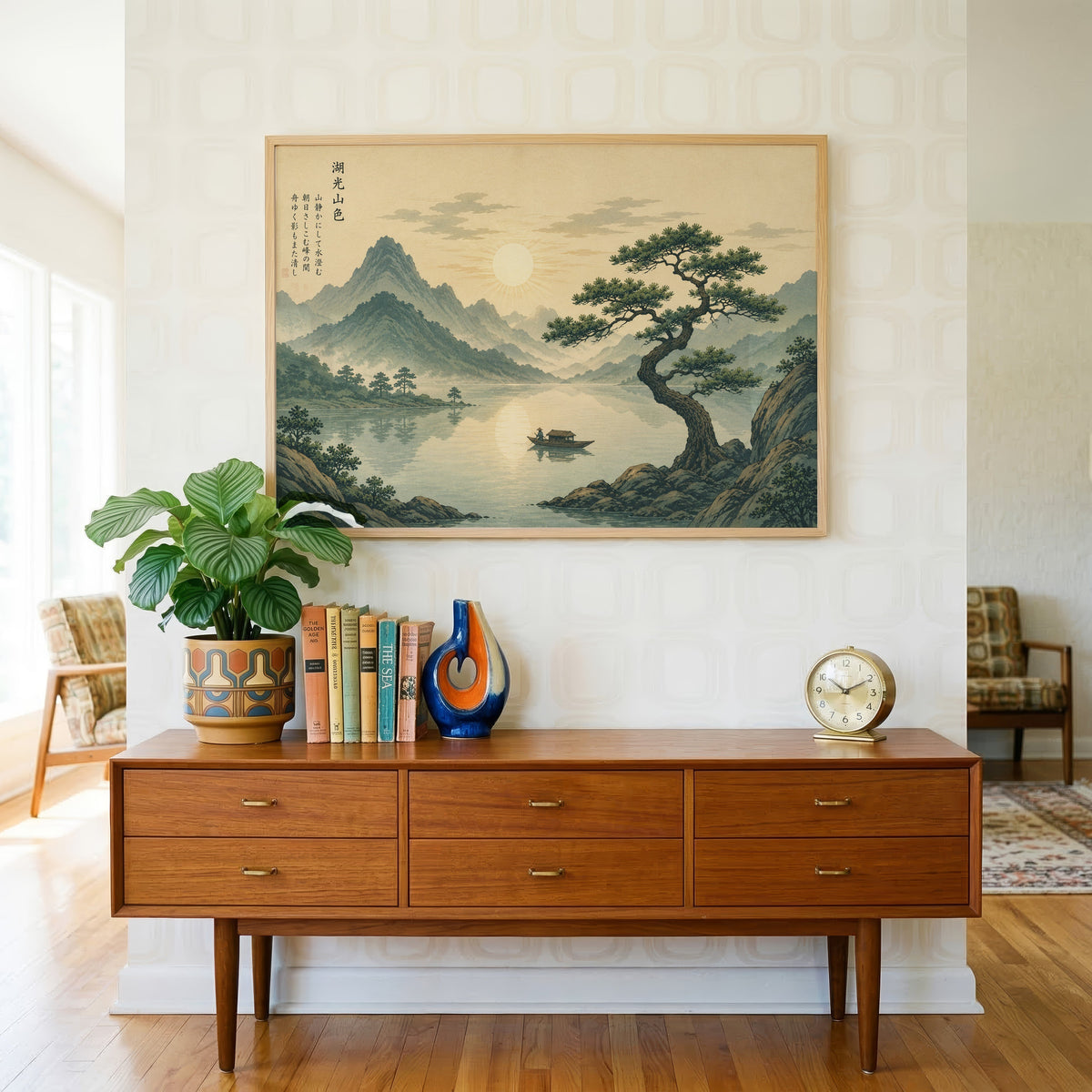 Pine tree over a mountain lake at sunrise print in a light oak frame above a teak credenza with books and a brass clock