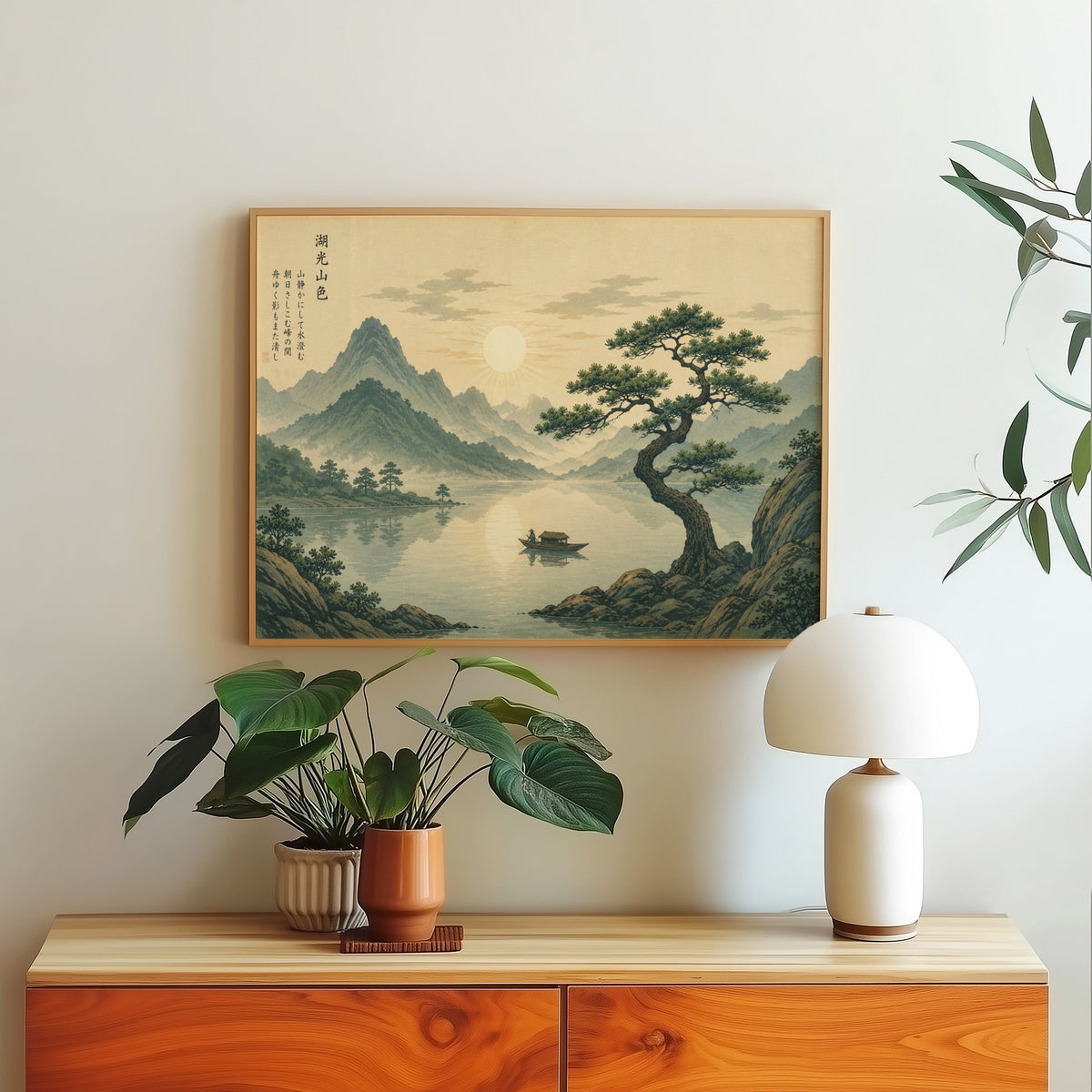 Pine tree over a mountain lake at sunrise print in a light oak frame above a wood sideboard with a white dome lamp