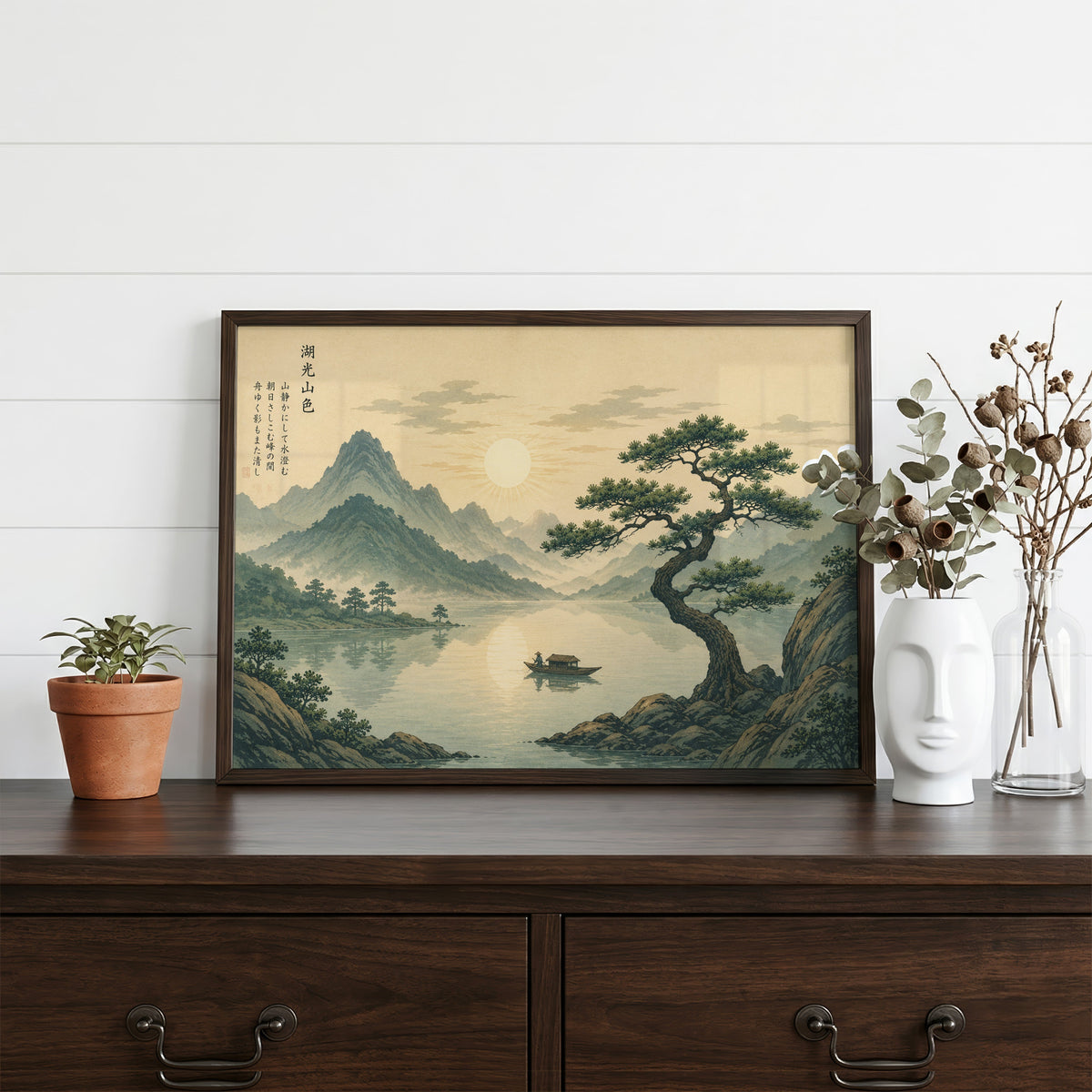 Pine tree over a mountain lake at sunrise print in a dark walnut frame on a dark wood dresser beside a white face vase