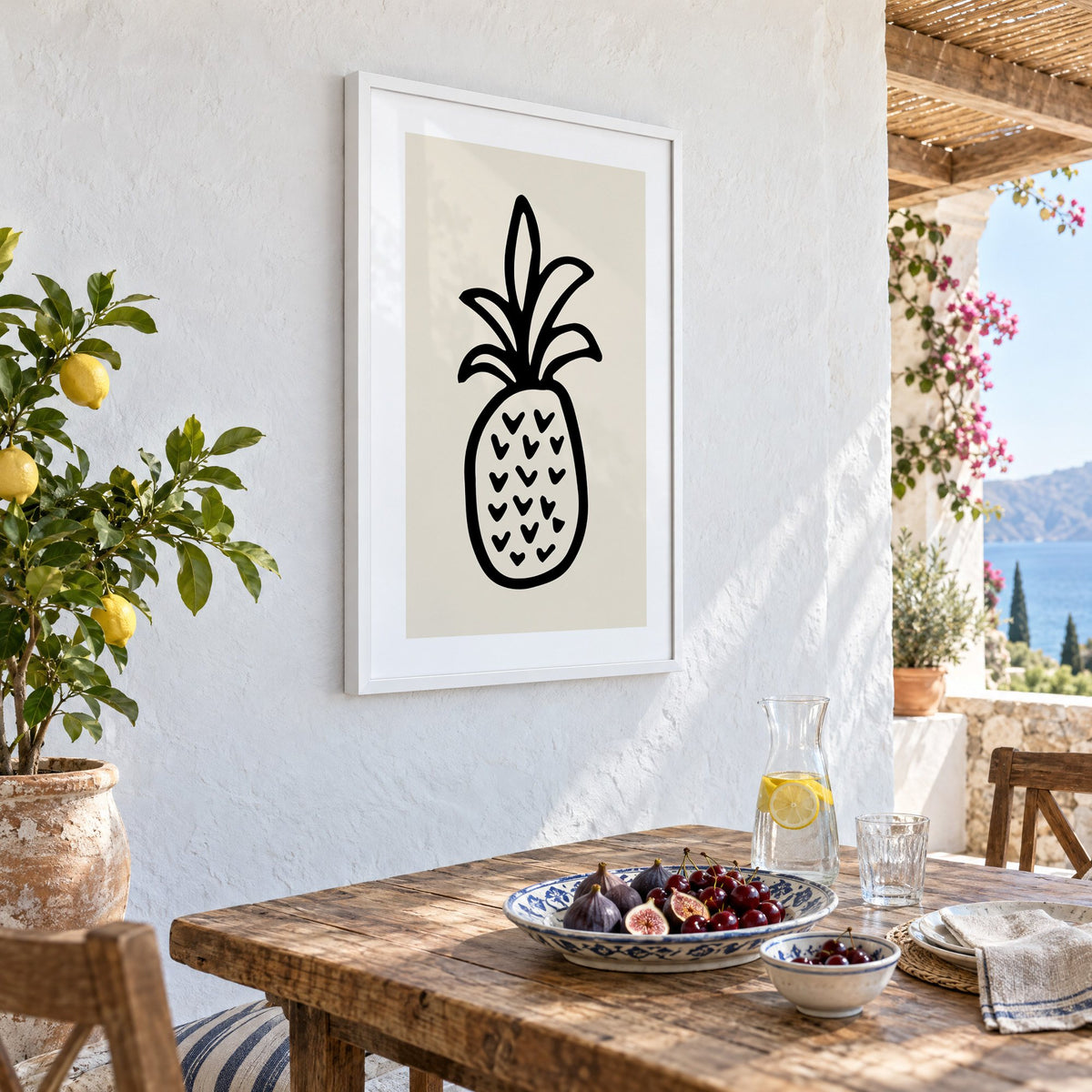 black ink line pineapple beige print in a white frame on a whitewashed terrace wall above a lunch table with figs and cherries, seen at an angle