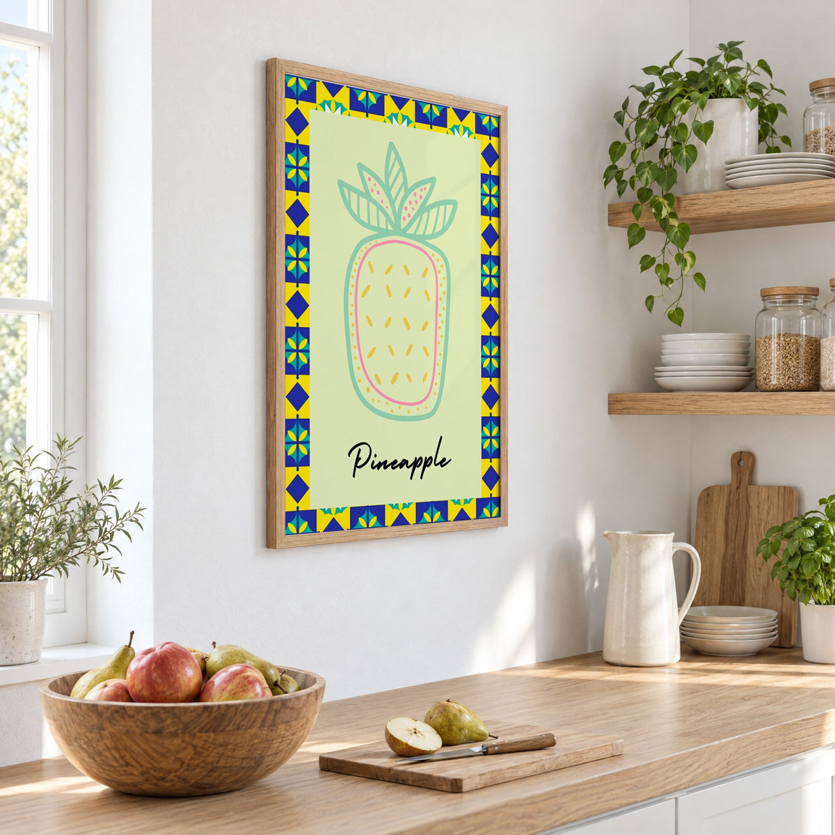 Pineapple mint line-art tile border print in an oak frame on a white kitchen wall above a counter with apples and pears, seen at an angle