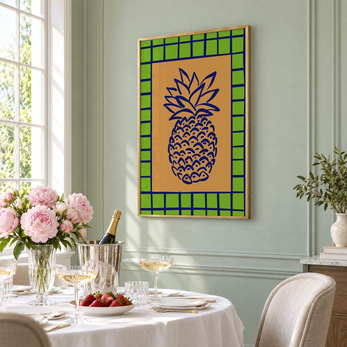 Pineapple navy line on ochre with green grid print in a brass frame on a sage wall above a brunch table with champagne and peonies, seen at an angle
