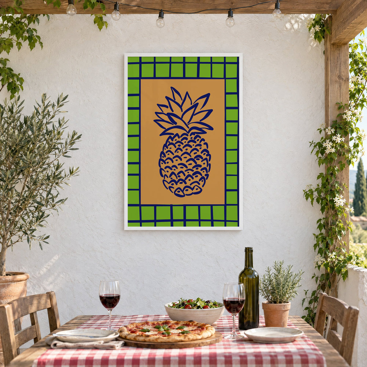 Pineapple navy line on ochre with green grid print in a white frame on a stucco wall above a checked-cloth table with pizza and red wine