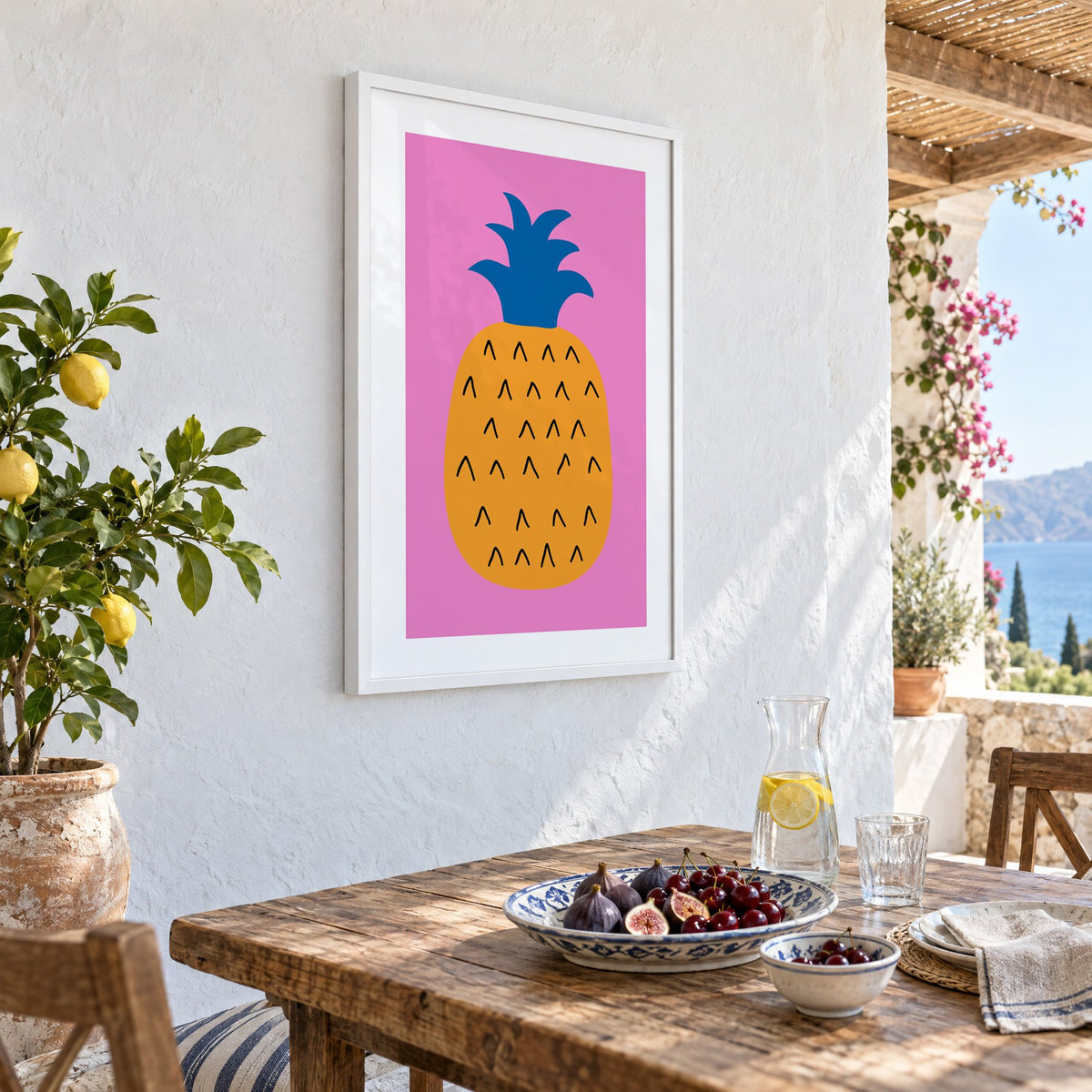 orange pineapple cobalt crown hot pink print in a white frame on a whitewashed terrace wall above a lunch table with figs and cherries, seen at an angle