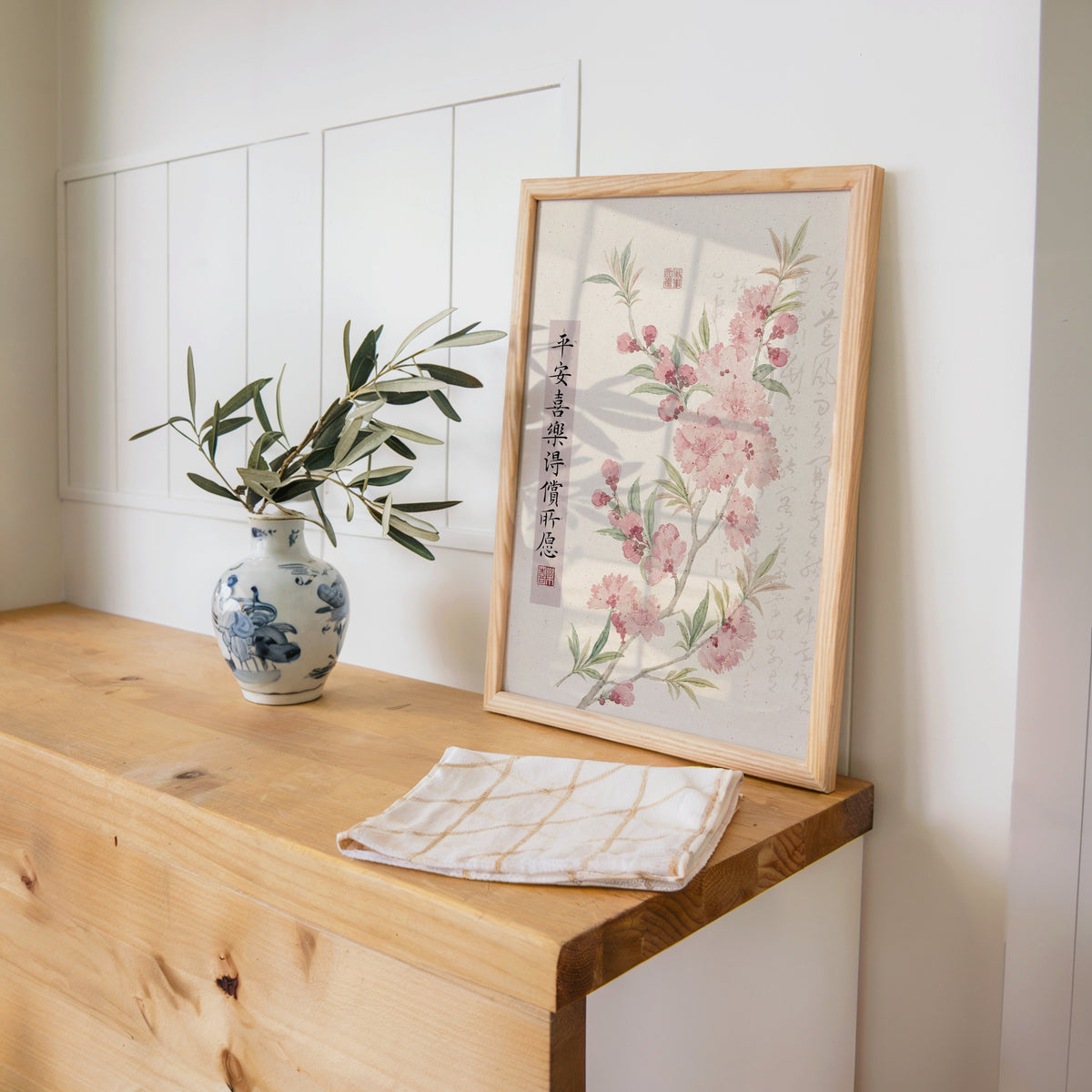 pink blossom with calligraphy blessing print in a pine frame on a wooden counter beside a blue and white porcelain vase