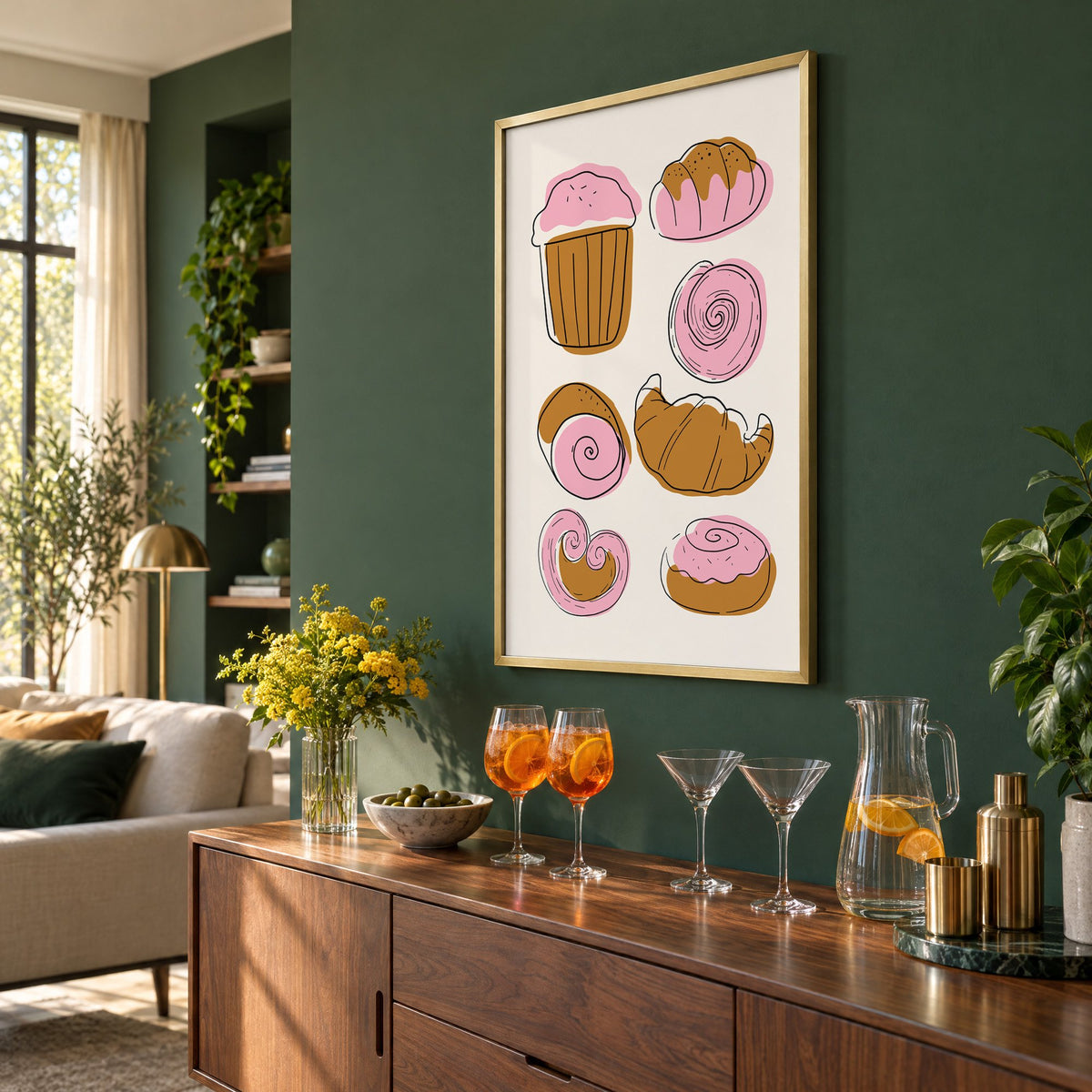 Pink and brown pastries doodle print in a brass frame on a bottle green wall above a sideboard with spritz and martini glasses, seen at an angle