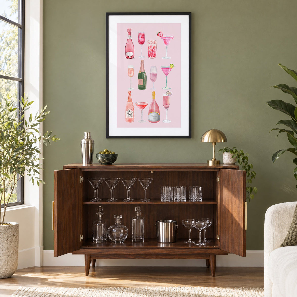 pink champagne bottles and cocktails watercolor print in a black frame on an olive green wall above a walnut bar cabinet with glassware