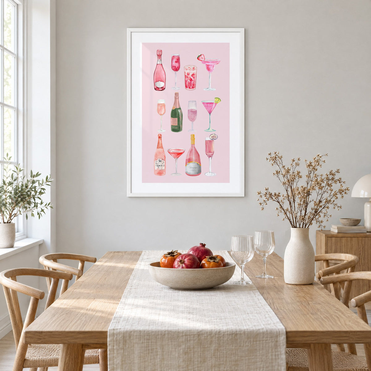 pink champagne bottles and cocktails watercolor print in a white frame above a light oak dining table with a linen runner and fruit bowl