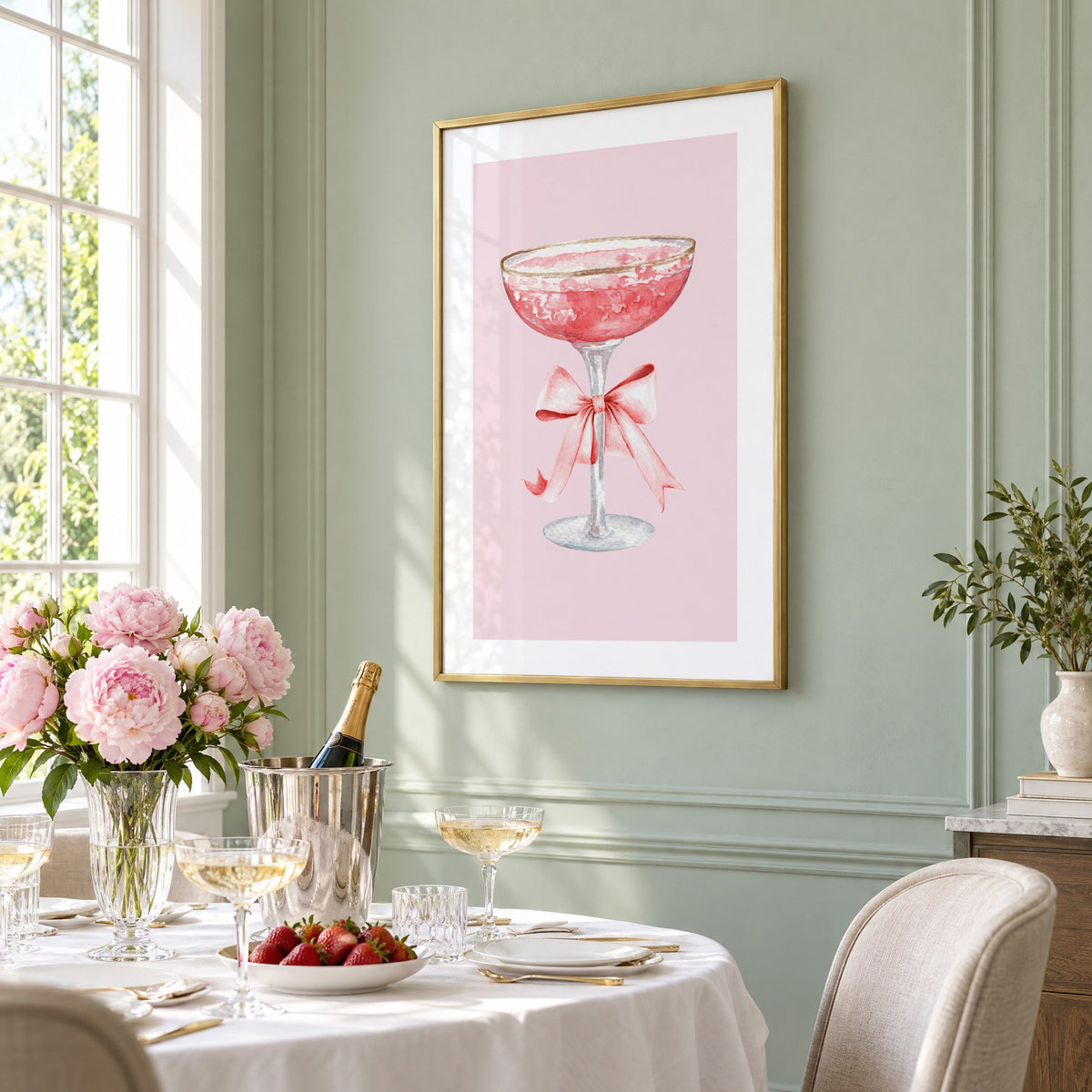gold rim champagne coupe satin bow print in a brass frame on a sage wall above a brunch table with champagne and peonies, seen at an angle