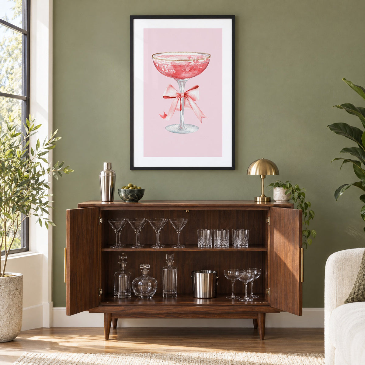 gold rim champagne coupe satin bow print in a black frame on an olive green wall above a walnut bar cabinet with glassware