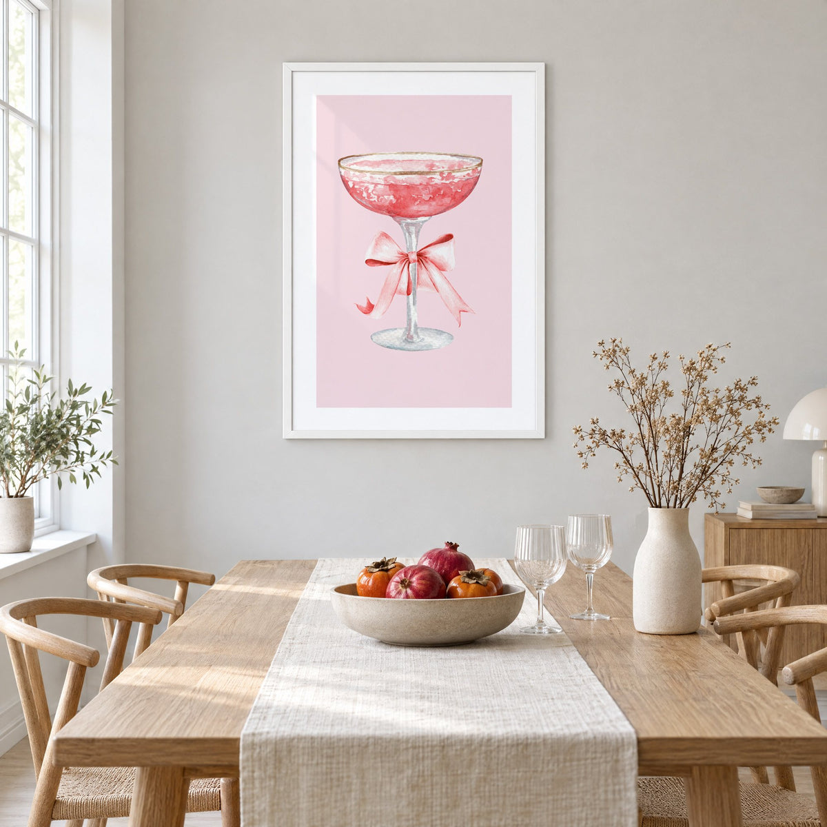 gold rim champagne coupe satin bow print in a white frame above a light oak dining table with a linen runner and fruit bowl