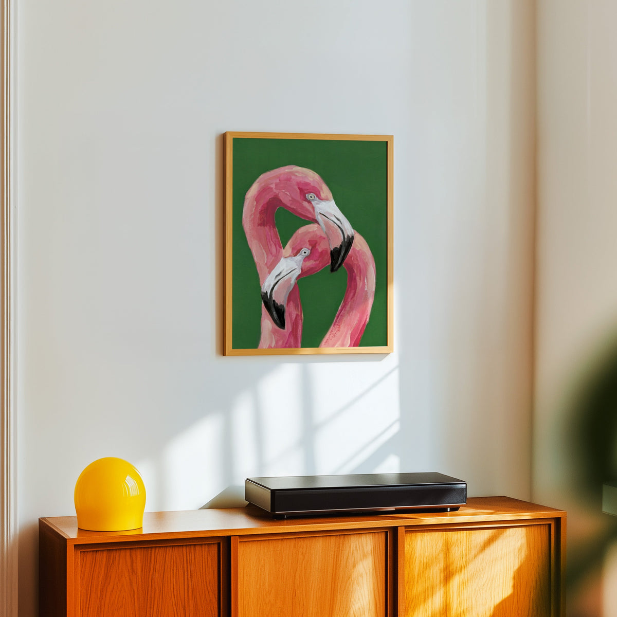Tropical flamingo painting print in a light oak frame above a teak sideboard with a yellow lamp