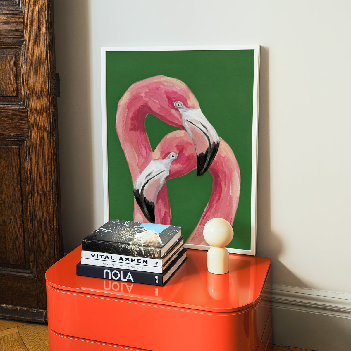 Two flamingos on green print in a white frame on a glossy orange cabinet with books