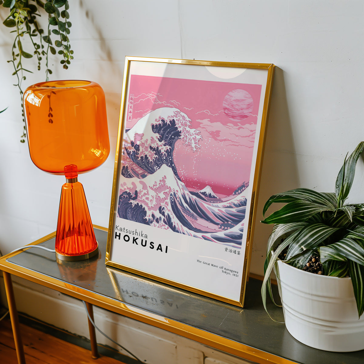 Pink Great Wave off Kanagawa print shown in a gold frame on a console beside an orange glass lamp