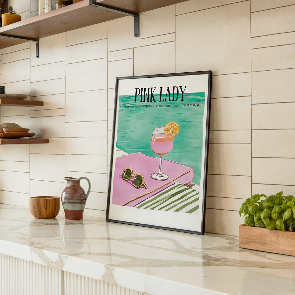 Pink Lady cocktail print in a black frame leaning on a marble counter in a tiled kitchen