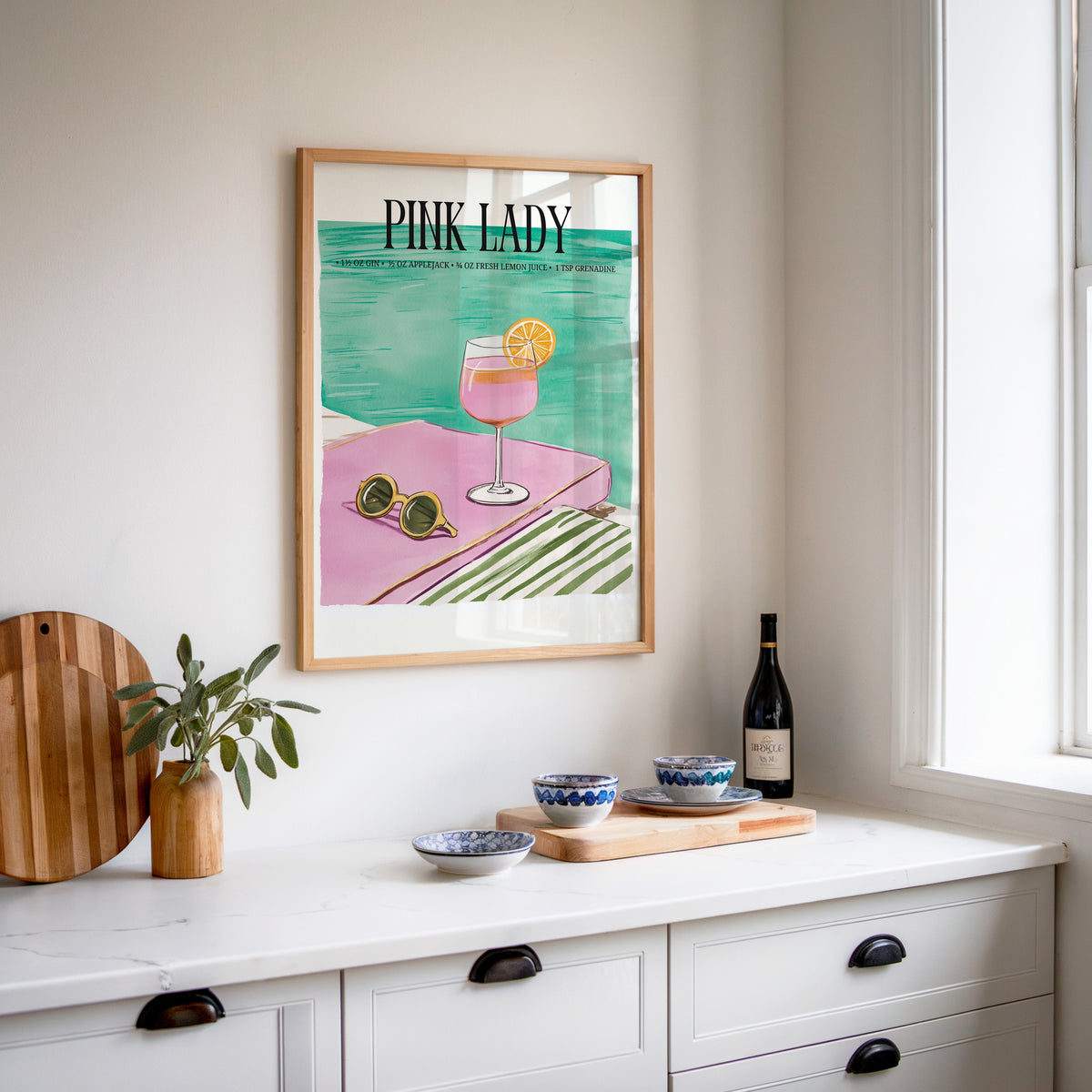 Pink Lady cocktail print in an oak frame above a white sideboard with a bottle of wine