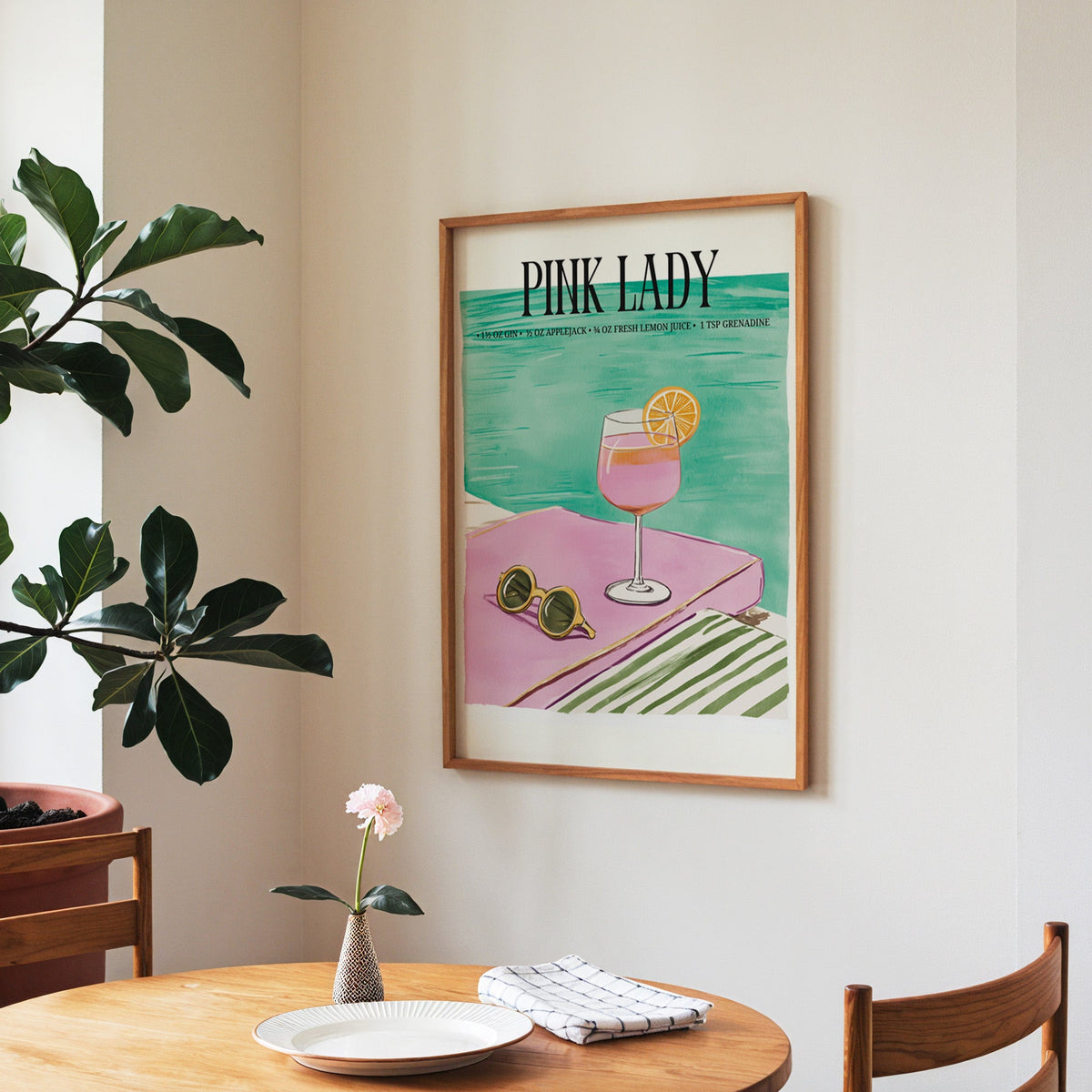 Pink Lady cocktail print in a walnut frame above a round table beside a fiddle-leaf fig