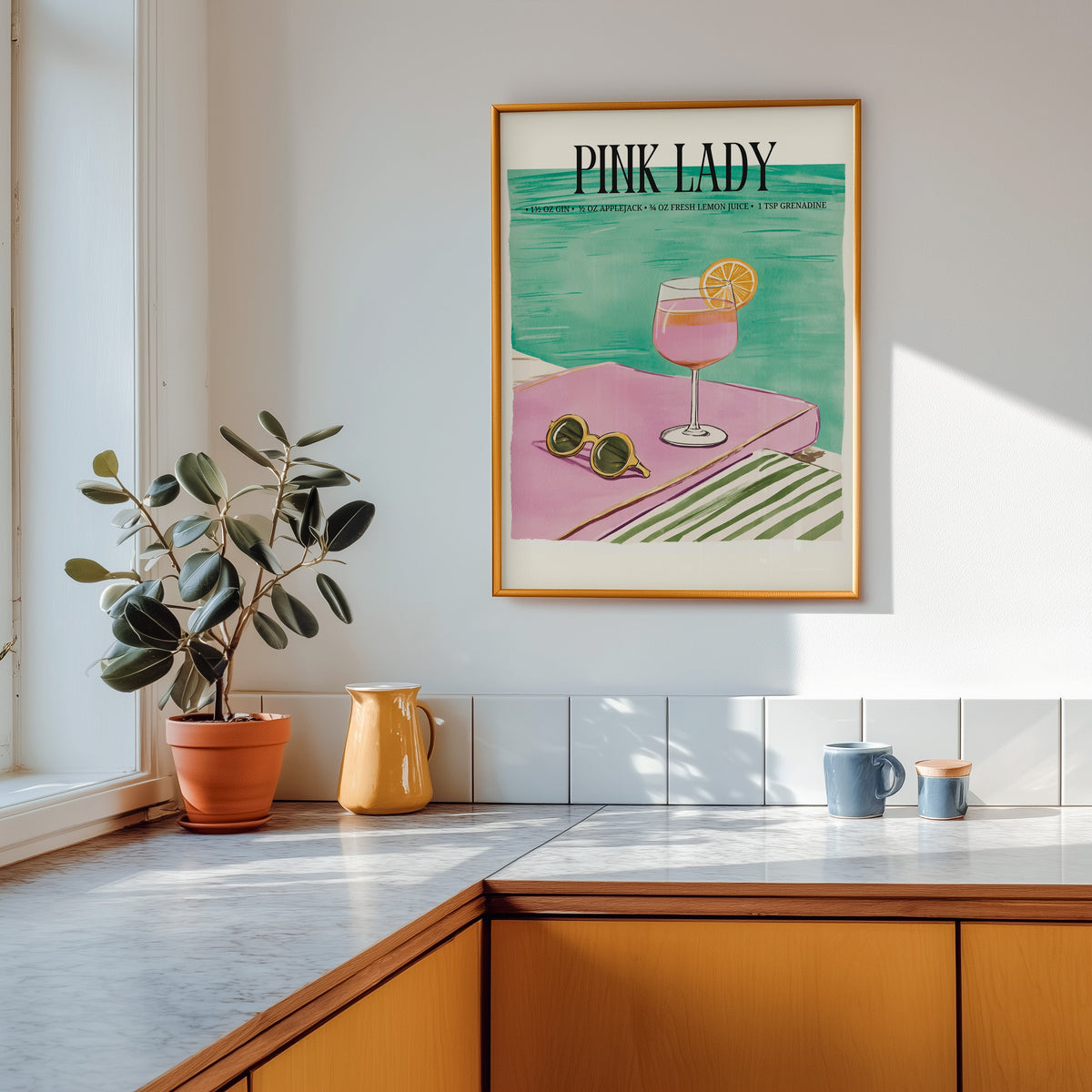 Pink Lady cocktail print shown in a gold frame above a marble counter in a tan kitchen