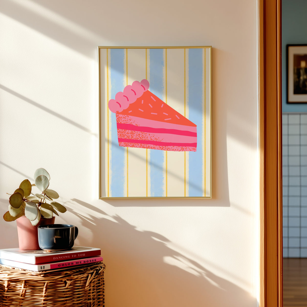 Pink layer cake print in a gold frame above a basket of books and a pilea plant