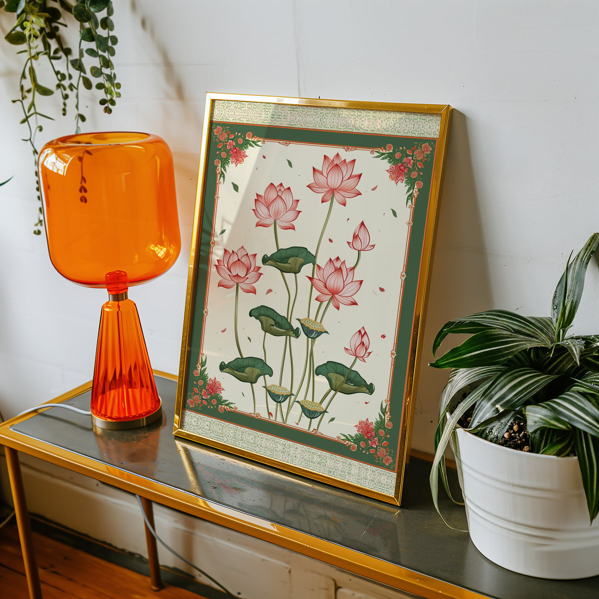 Pink lotus with green ornamental border print shown in a gold frame on a console beside an orange glass lamp