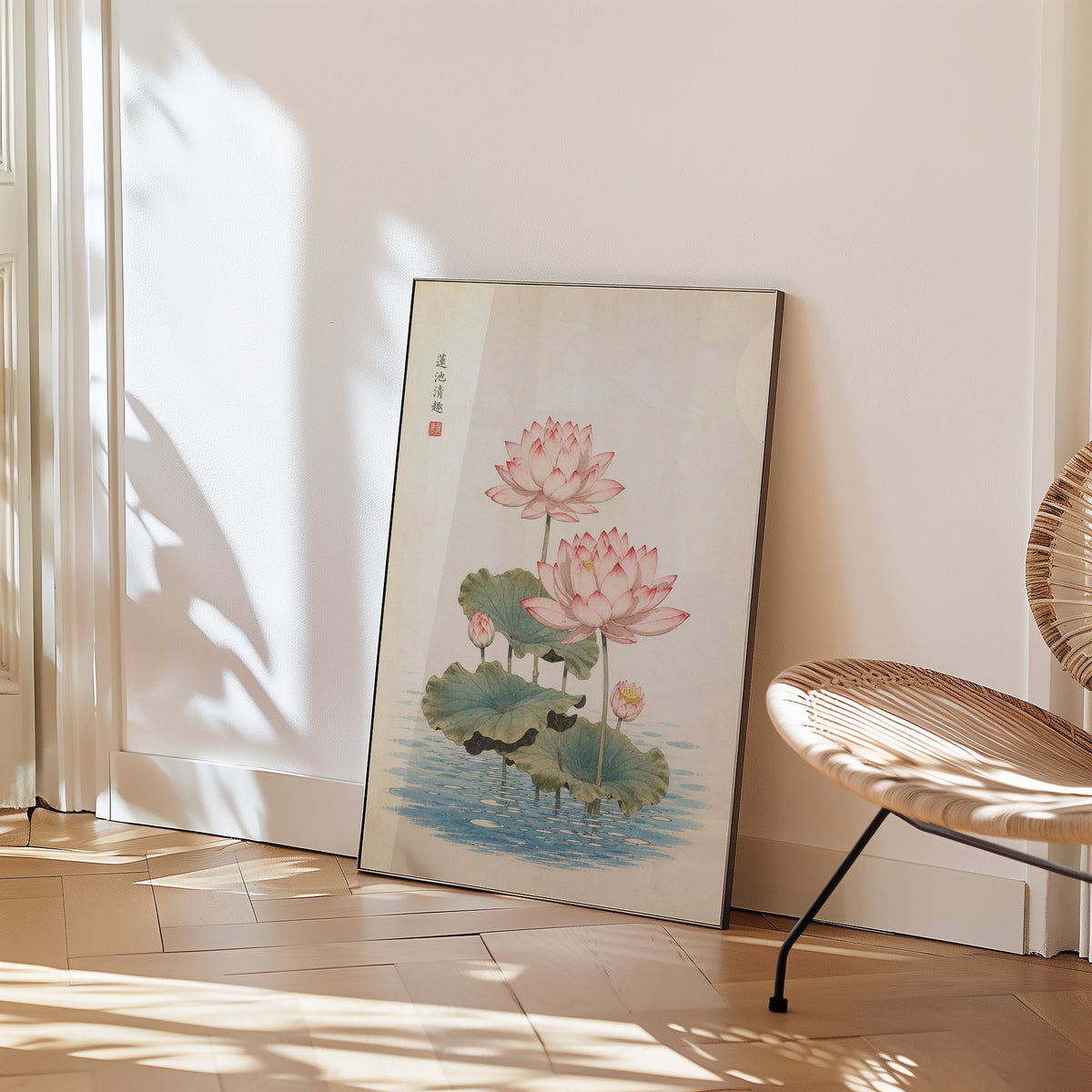 pink lotus pond print in a black frame leaning on a herringbone floor beside a woven rattan chair