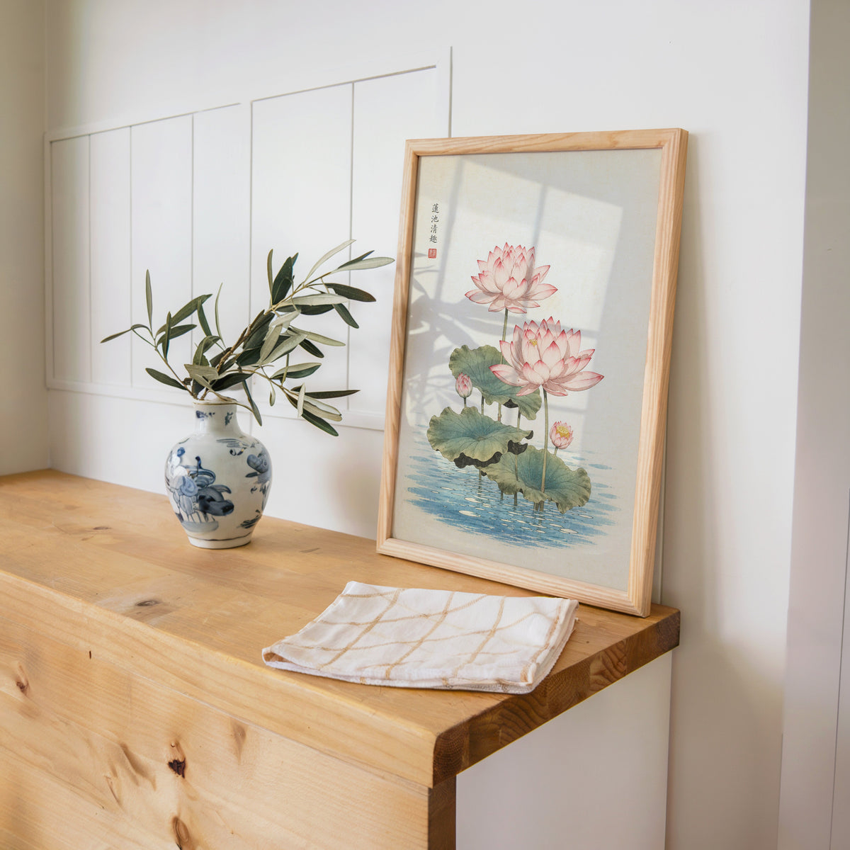 pink lotus pond print in a pine frame on a wooden counter beside a blue and white porcelain vase
