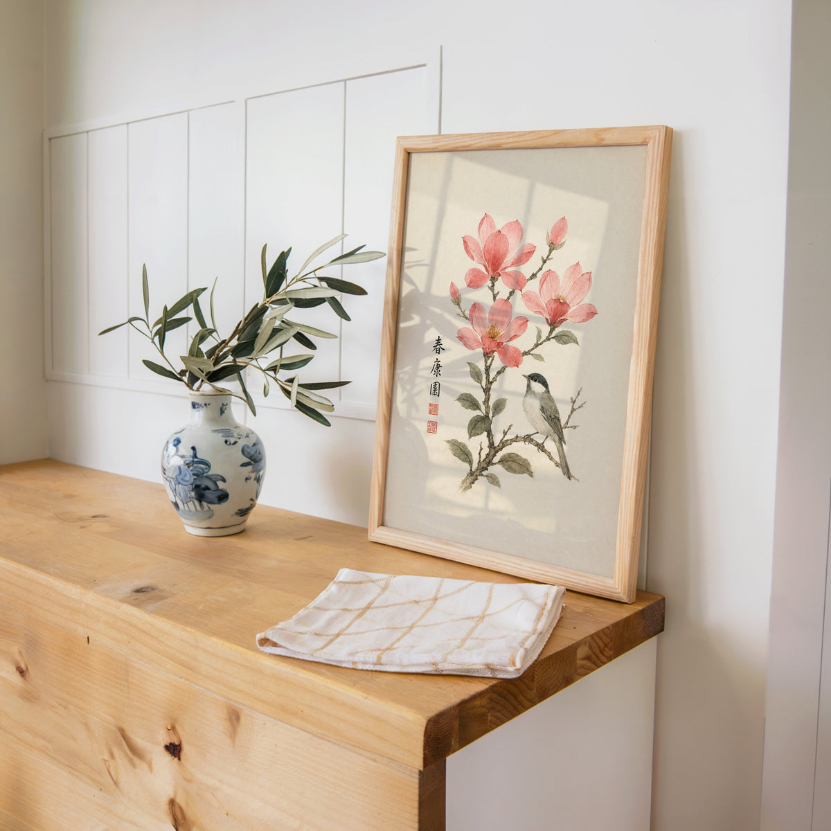 songbird on pink blossom branch print in a pine frame on a wooden counter beside a blue and white porcelain vase