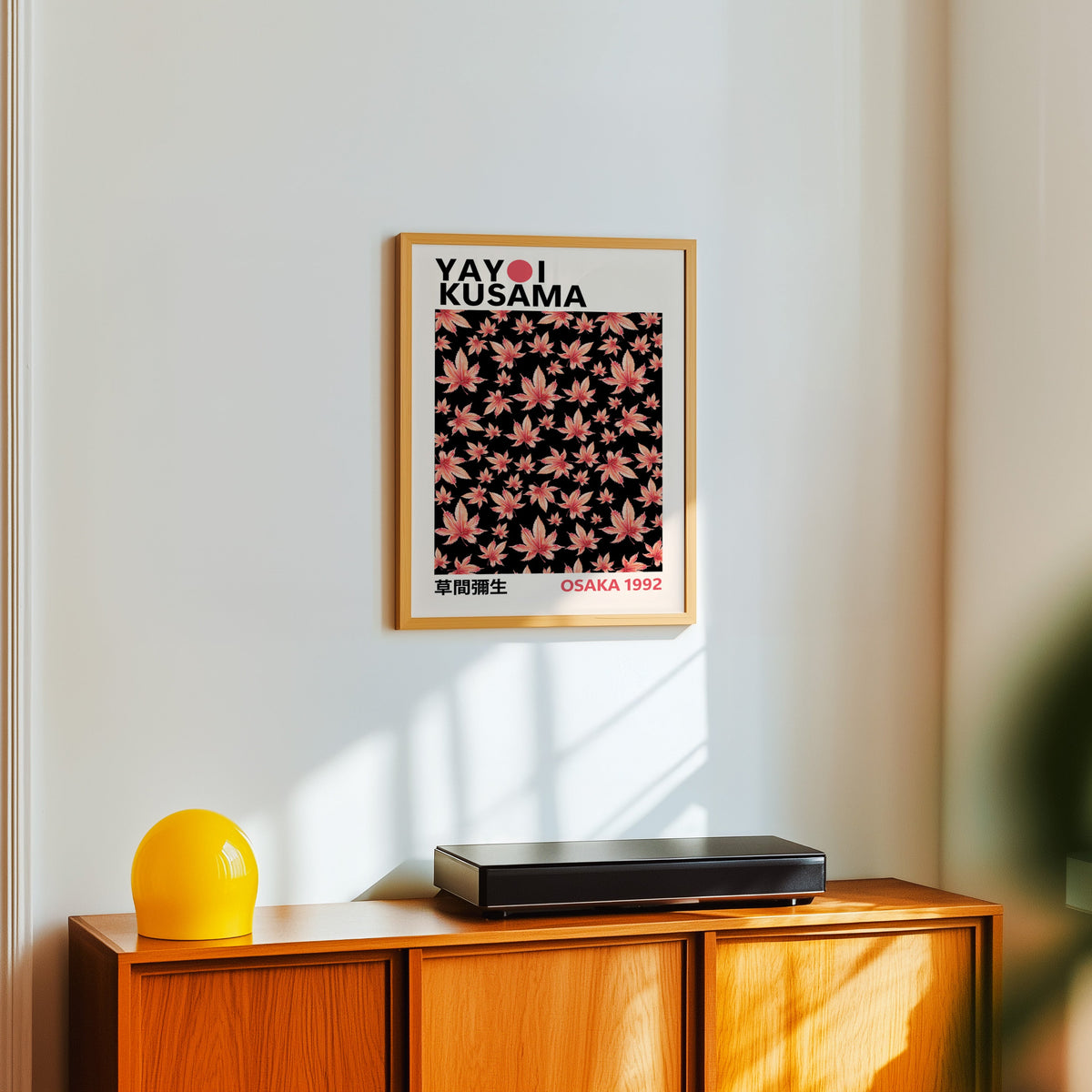 Yayoi Kusama inspired pink maple leaves on black print in an oak frame over a teak sideboard with a yellow lamp