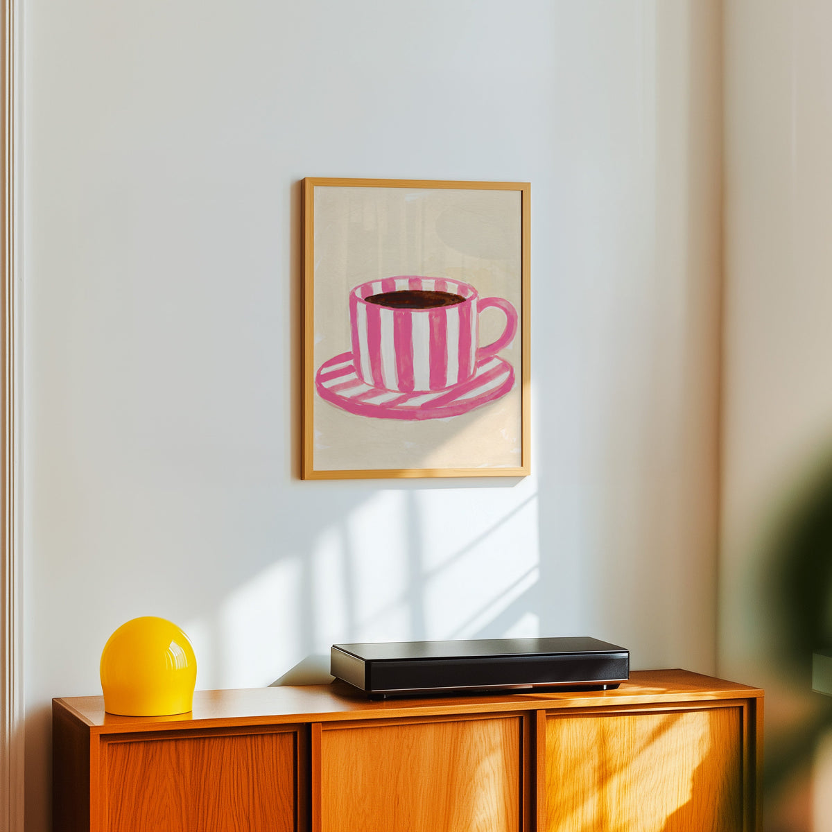 Pink coffee wall art print in a light oak frame above a teak sideboard with a yellow lamp